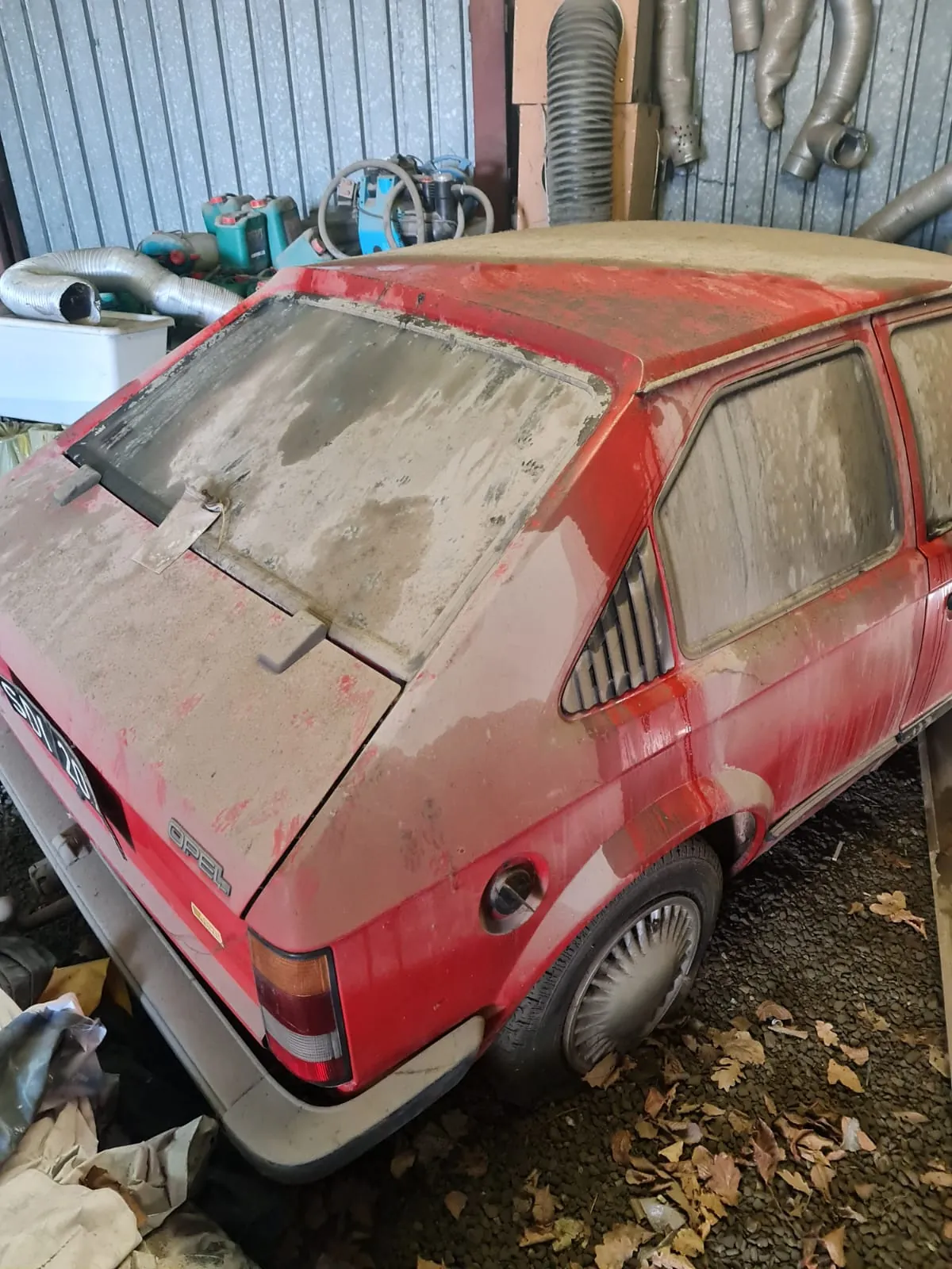 Czerwony samochód marki Opel pokryty kurzem i brudem, widok od tyłu. Na tylnej szybie i bagażniku widoczne ślady zabrudzeń. W tle warsztat z metalową ścianą, przewodami, zbiornikami i innymi narzędziami. Na ziemi żwir i opadłe liście. Na bagażniku widoczny napis OPEL oraz tablica rejestracyjna po lewej stronie.
