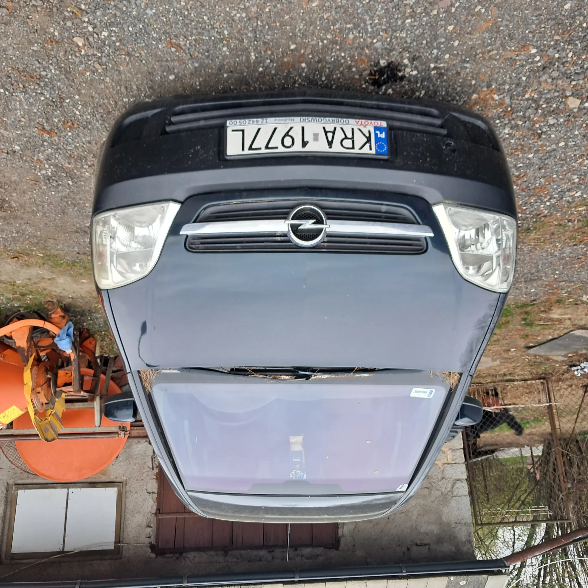 Ciemnoszary samochód marki Opel z przodu, z tablicą rejestracyjną KRA 1977L. Na tablicy widoczne dodatkowe napisy: PL, TOYOTA DOBRYGOWSKI oraz numer 124420500. W tle budynek z oknem i bramą, pomarańczowe maszyny rolnicze, żwirowa powierzchnia oraz ogrodzenie. Na przedniej szybie samochodu znajdują się naklejki.