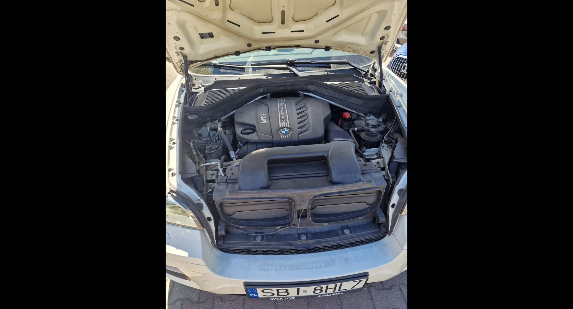 Widok z góry na otwartą maskę białego samochodu marki BMW, ukazującą silnik i jego komponenty. Na dole zdjęcia widoczna jest tablica rejestracyjna o treści 'SB I 8HL7' z oznaczeniem 'PL' oraz napisem 'WEKTOR' i 'Białystok-Biała' na ramce.