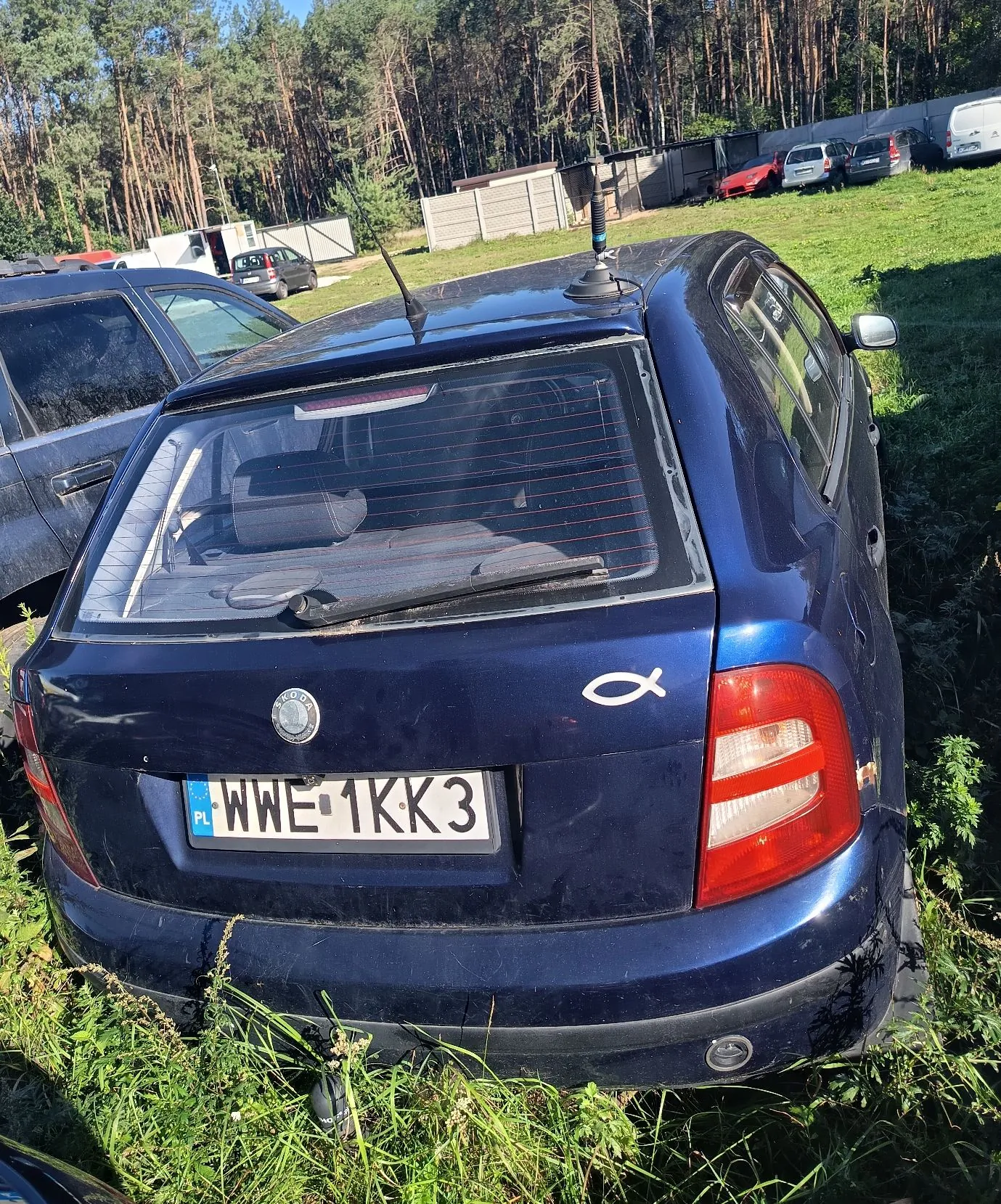 Tylny widok ciemnoniebieskiego samochodu marki Skoda zaparkowanego na trawiastym terenie. Samochód ma polską tablicę rejestracyjną o numerze WWE 1KK3. Na klapie bagażnika, po prawej stronie, znajduje się biały symbol nieskończoności. W tle widoczny jest las, betonowy płot oraz inne zaparkowane samochody.