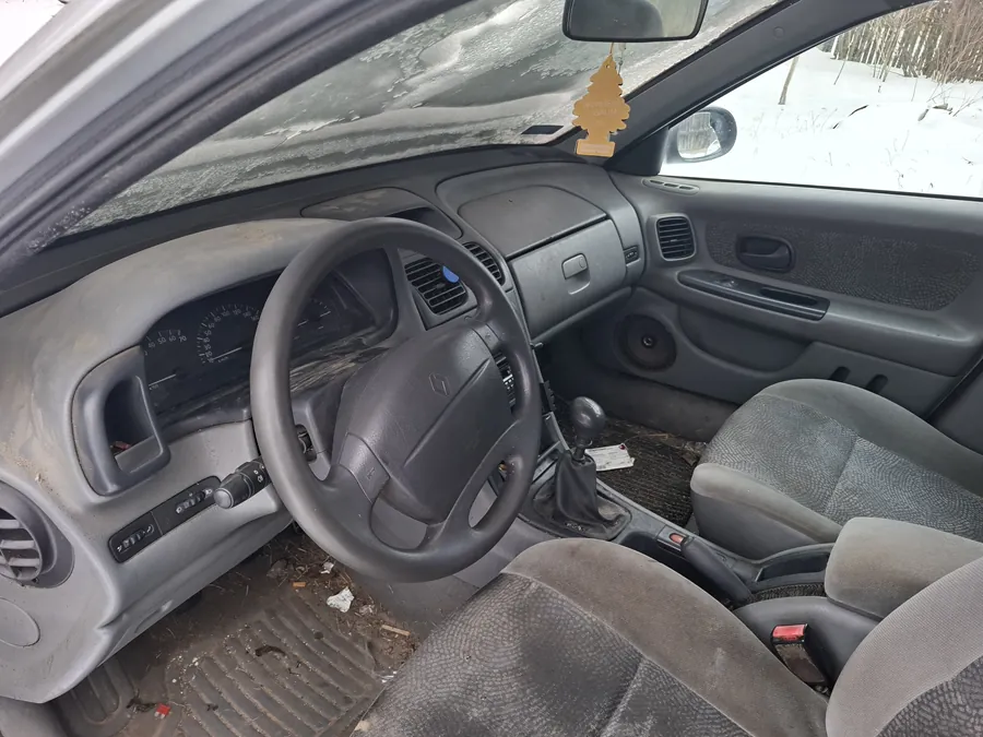 Wnętrze samochodu z widocznym kierownicą, deską rozdzielczą, siedzeniami i przekładnią. Kierownica po lewej stronie, na lusterku wstecznym wiszący żółty odświeżacz powietrza w kształcie drzewka. Deska rozdzielcza i siedzenia są zabrudzone, na podłodze widoczne śmieci. Poza samochodem śnieg i drzewa. Wnętrze wydaje się nieczyste i zużyte.