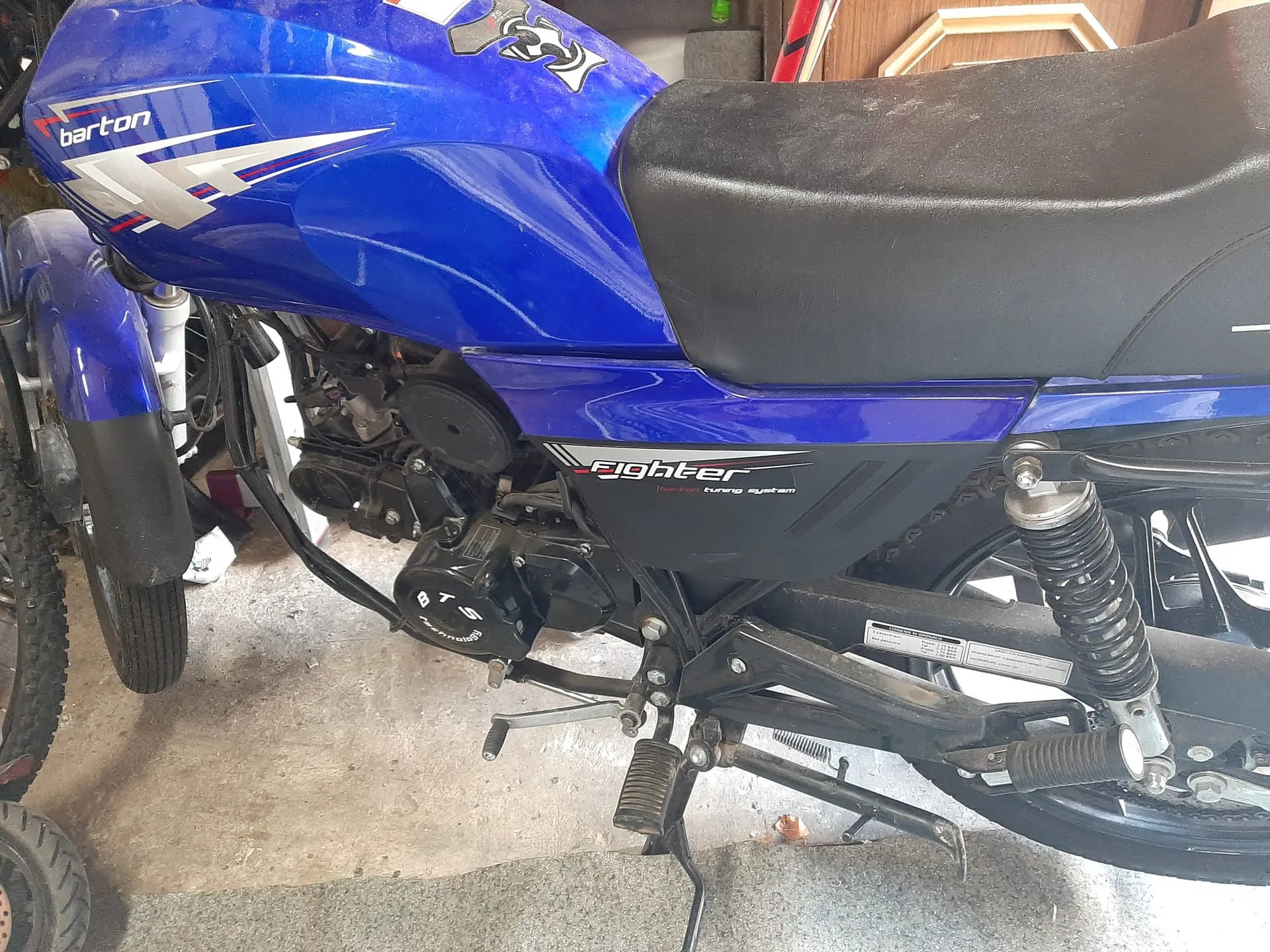 Niebieski motocykl marki Barton z czarnym siedzeniem. Na baku widoczny napis 'barton' oraz grafika z białymi i czerwonymi elementami. Na bocznej części kadłuba napis 'fighter' oraz 'tuning system'. Widoczny czarny silnik z napisem 'BTS'. W tle betonowa podłoga, fragmenty innych pojazdów (koła) oraz drewniane drzwi. Na ramie motocykla znajduje się biała tabliczka z czarnym tekstem.