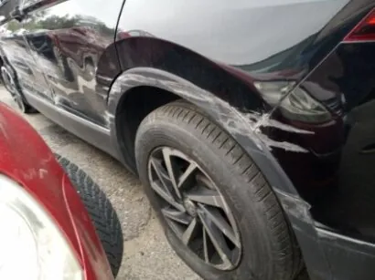 Czarny samochód z widocznymi białymi zadrapaniami na boku, w okolicy tylnego koła. Część czerwonego pojazdu i asfaltowa powierzchnia na pierwszym planie.