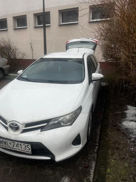 Biały samochód marki Toyota z otwartym bagażnikiem zaparkowany przy budynku. Widoczna tablica rejestracyjna WWL 74135. W tle budynek z oknami, krzewy i śnieg na ziemi. Częściowo widoczny inny samochód po lewej stronie.
