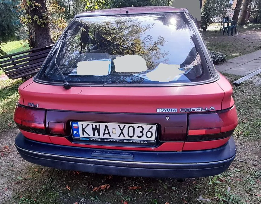 Tylny widok czerwonego samochodu Toyota Corolla w wersji XLI. Pojazd posiada polską tablicę rejestracyjną o numerze KWA X036. Na tylnej szybie widoczne są białe kartki papieru. Samochód stoi zaparkowany na trawiastym terenie obok drewnianej ławki.