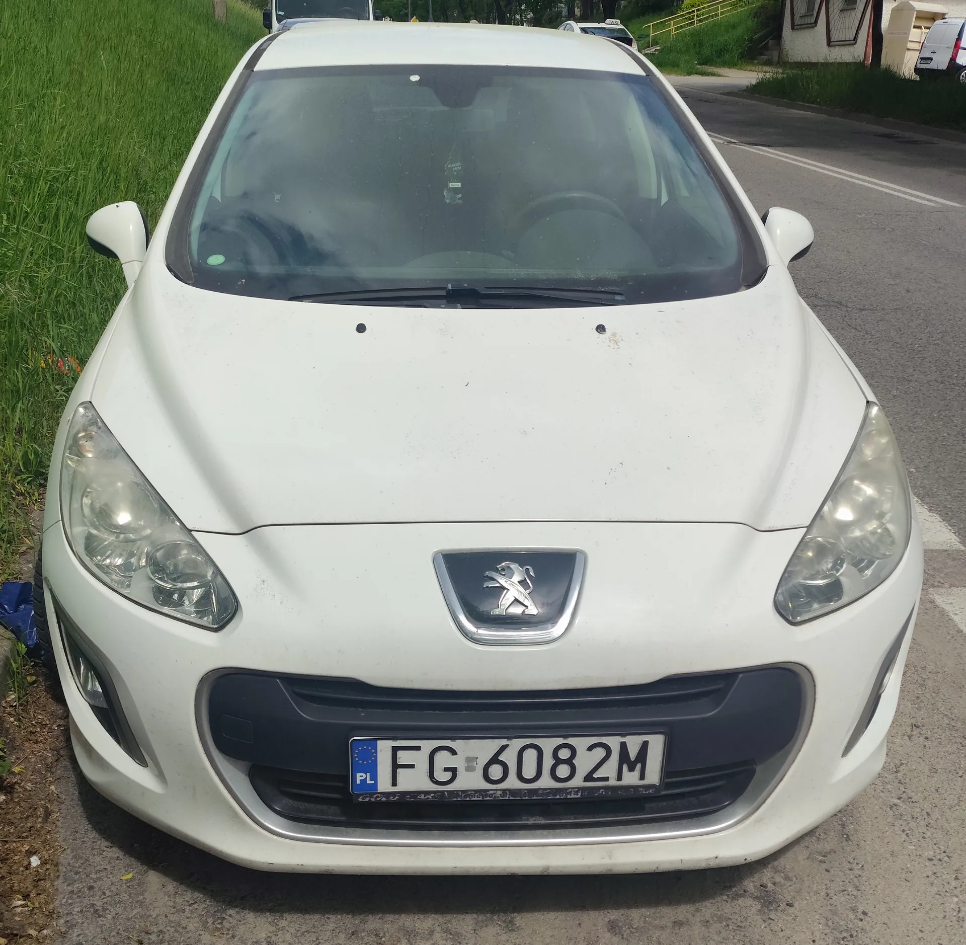 Biały samochód marki Peugeot zaparkowany na poboczu drogi. Widoczna przód pojazdu z logo marki na masce, tablicą rejestracyjną FG-6082M z niebieskim pasem PL po lewej stronie. Po lewej stronie samochodu trawa, po prawej asfaltowa droga z białą linią. W tle inne pojazdy, zielone krzewy i fragment budynku z żółtymi schodami.