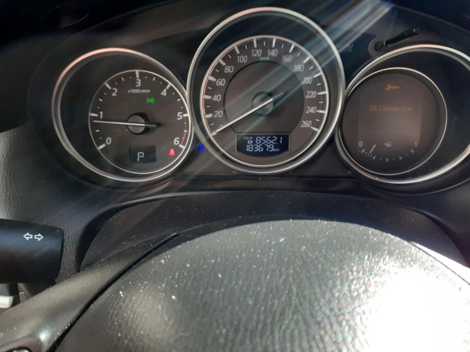 Zbliżenie na deskę rozdzielczą samochodu z trzema okrągłymi wskaźnikami. Centralny wskaźnik to prędkościomierz z zakresem do 260 km/h, pokazujący aktualnie 0 km/h, z cyfrowym wyświetlaczem przebiegu TRIP 8562.1 i 183679 km. Po lewej stronie znajduje się obrotomierz z oznaczeniem x1000r/min oraz zapaloną czerwoną kontrolką parkowania (litera P). Po prawej stronie znajduje się wyświetlacz z napisem 'Oil Change Due' oraz pomarańczową ikoną olejarki.