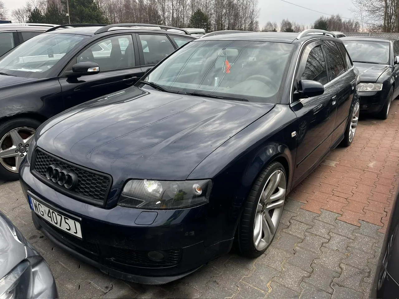 Ciemnoniebieski samochód Audi kombi z tablicą rejestracyjną NG 4075 J zaparkowany na placu parkingowym. W tle inne pojazdy, drzewa i pochmurne niebo. Samochód posiada czarną atrapę chłodnicy z czterema pierścieniami marki, srebrne felgi oraz dachowy bagażnik. Podłoga wyłożona czerwonymi i szarymi kostkami brukowymi.