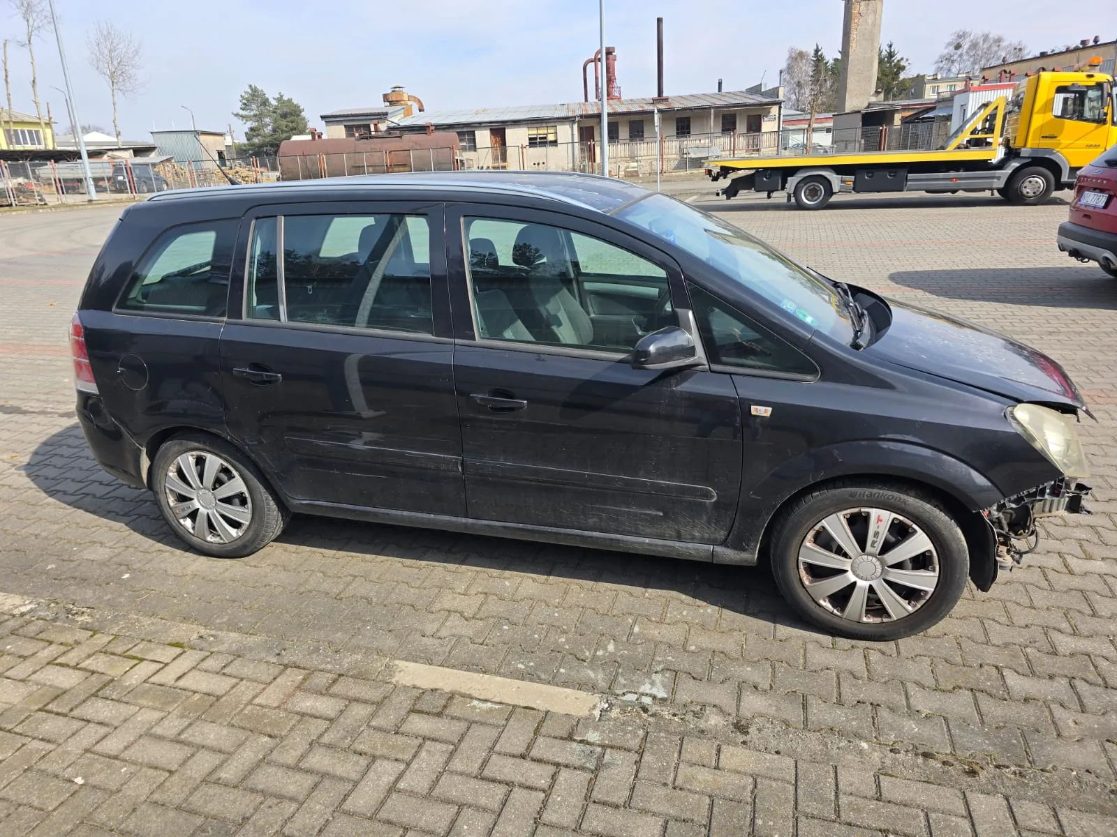 Czarny samochód typu minivan z uszkodzonym przodem, brakującym zderzakiem i uszkodzonym reflektorem. Pojazd stoi na brukowanej powierzchni. W tle widoczne są przemysłowe budynki, żółty holownik oraz czerwony samochód. Niebo jest jasne, a na tle znajdują się również drzewa i kominy.