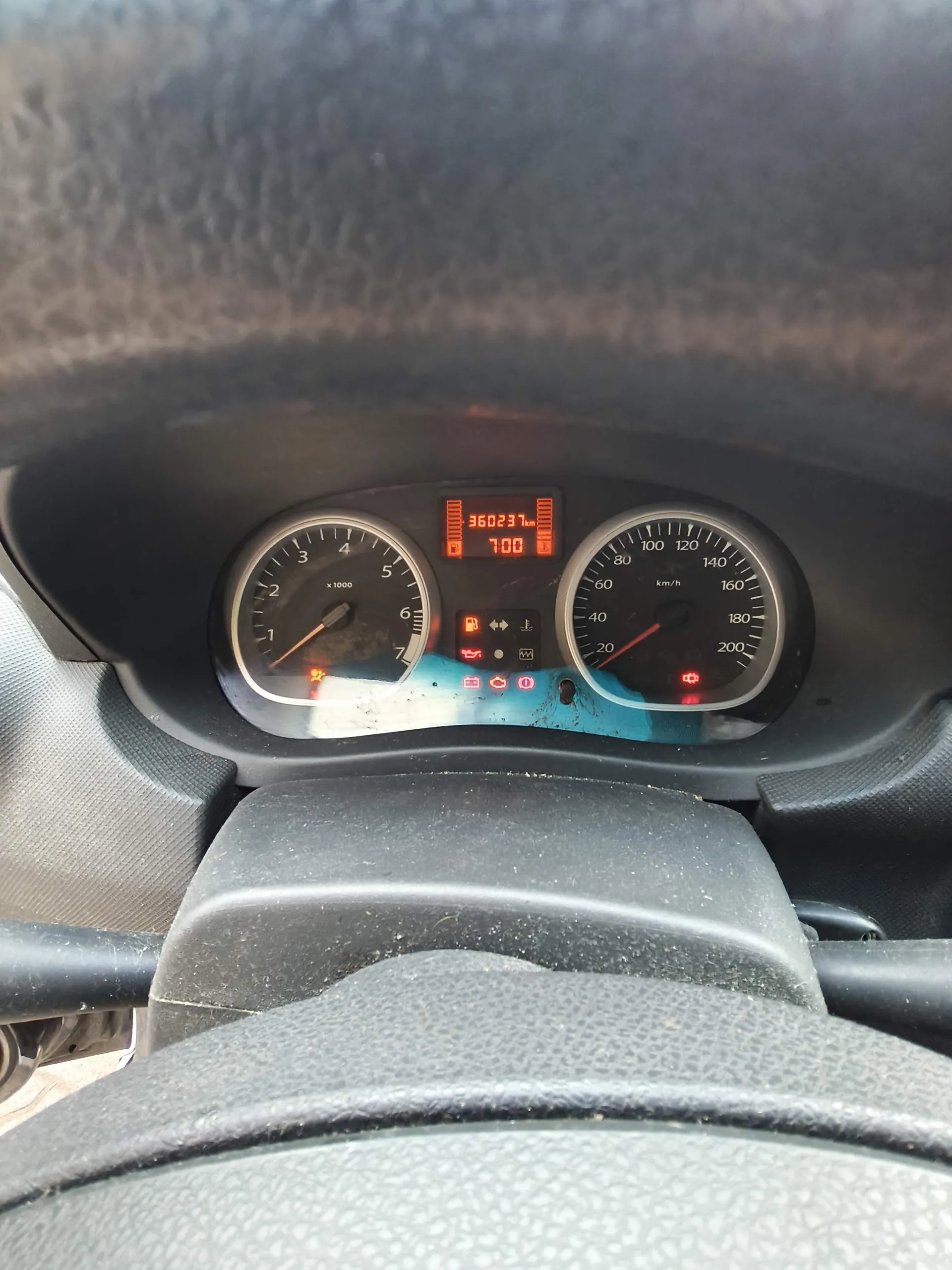 Widok z perspektywy kierowcy na deskę rozdzielczą samochodu. W centrum znajduje się cyfrowy licznik przebiegu wskazujący 360237 km oraz zegar z godziną 7:00. Po lewej stronie znajduje się analogowy obrotomierz z podziałką od 1 do 7 x 1000 obr./min, a po prawej analogowy prędkościomierz z podziałką do 200 km/h. Pomiędzy nimi widoczne są kontrolki ostrzegawcze, w tym zapalona czerwona kontrolka akumulatora oraz kontrolka oleju. W tle widoczna jest czarna, teksturowana obudowa deski rozdzielczej oraz fragment kierownicy na dole obrazu.