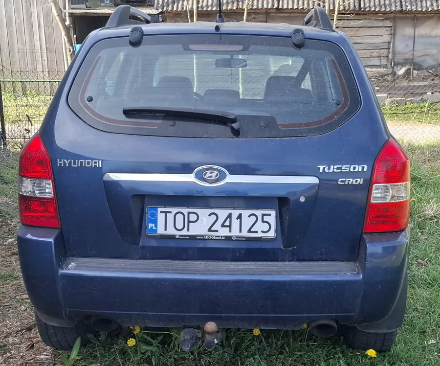 Tył niebieskiego samochodu Hyundai Tucson CRDi z tablicą rejestracyjną TOP 24125 (PL). Widoczne są czerwone światła tylnego, logo marki, napisy HYUNDAI po lewej stronie i TUCSON CRDi po prawej. Samochód stoi na trawie z żółtymi kwiatami, na tle drewnianego budynku i ogrodzenia. Tekst na tablicy rejestracyjnej jest czytelny i poziomy.