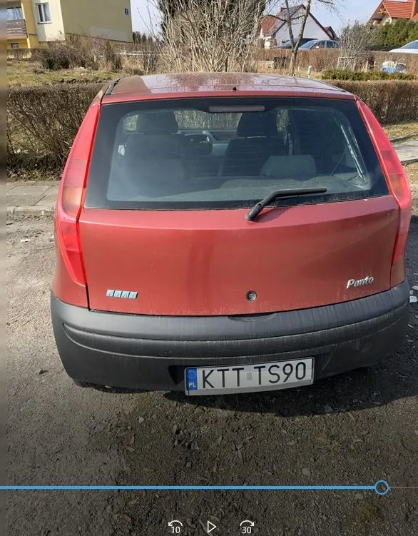 Czerwony samochód Fiat Punto widziany od tyłu z tablicą rejestracyjną KTT TS90 (PL) na środku tylnego zderzaka. Na bagażniku widoczne napisy "Fiat" po lewej i "Punto" po prawej stronie. Tylne okno z wycieraczką. Samochód zaparkowany na żwirowym podjeździe, na tle domów mieszkalnych, krzewów i jasnego nieba.