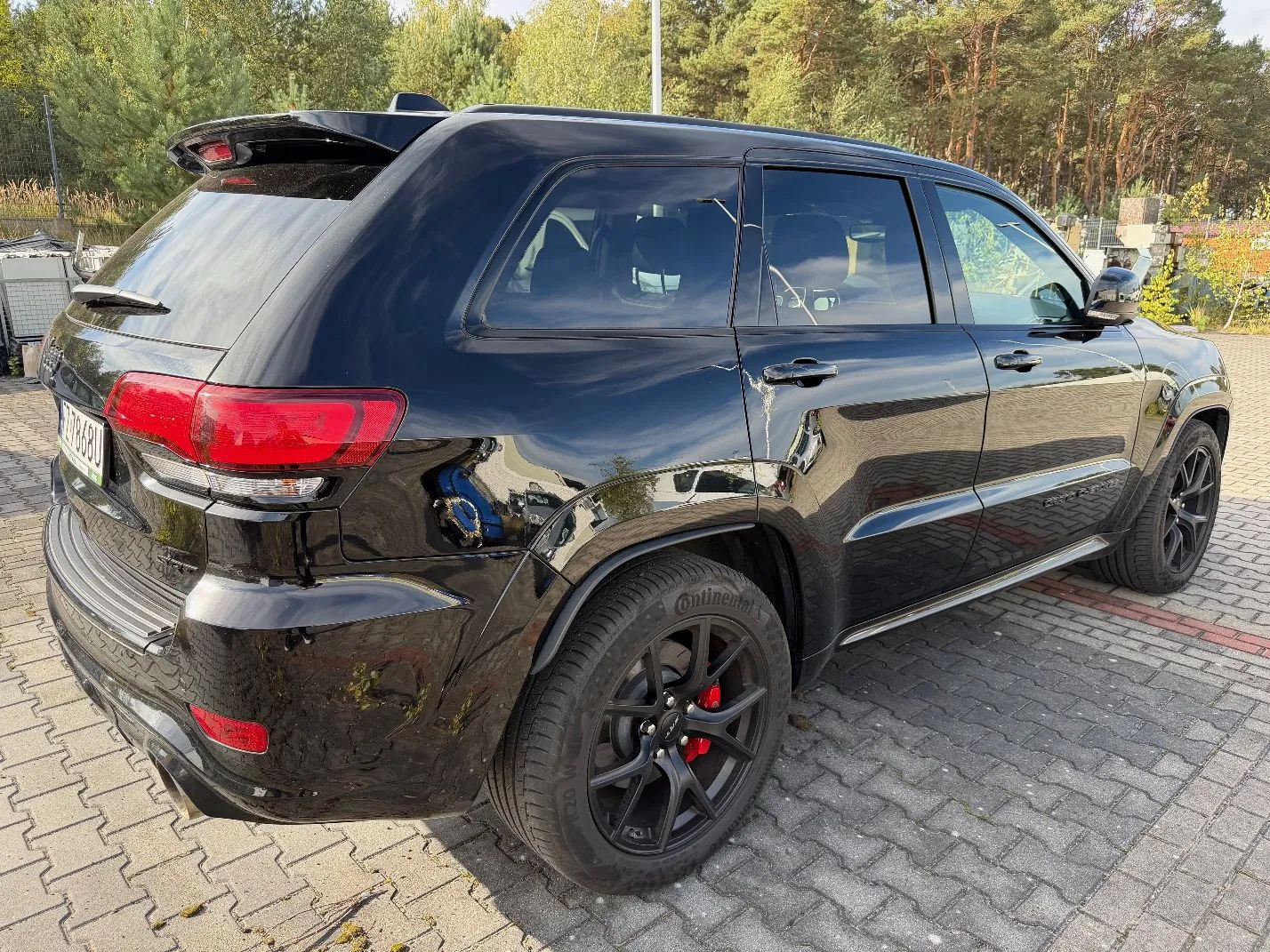 Czarny samochód typu SUV zaparkowany na kostce brukowej. Pojazd jest widoczny z tyłu i z boku, posiada przyciemniane szyby, czarne felgi z czerwonymi zaciskami hamulcowymi oraz spoiler na dachu. W tle widoczne są zielone drzewa i las.
