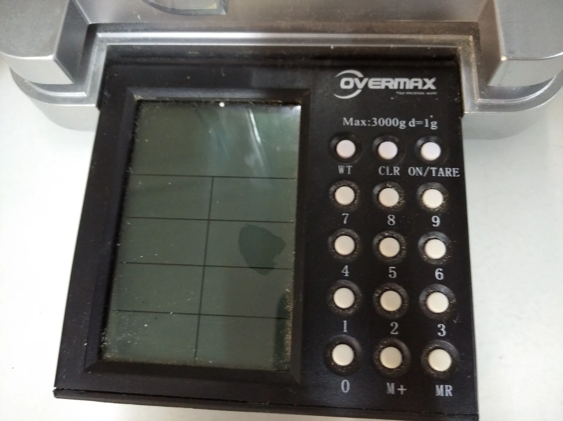Zbliżenie na panel sterowania wagi elektronicznej marki OVERMAX. Urządzenie posiada prostokątny wyświetlacz LCD po lewej stronie oraz panel z białymi przyciskami po prawej. Nad przyciskami znajduje się napis 'Max:3000g d=1g'. Przyciski są opisane jako: WT, CLR, ON/TARE, cyfry od 0 do 9, M+ oraz MR.