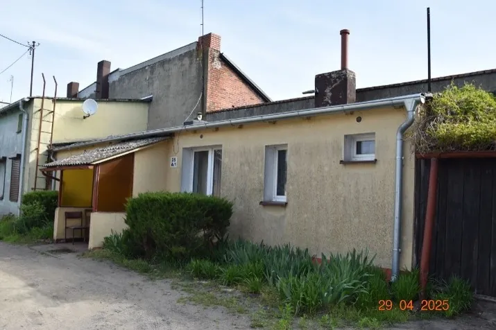 Zdjęcie niskiego, parterowego budynku o żółtej elewacji z płaskim dachem i rynną. W ścianie znajdują się dwa okna oraz drzwi wejściowe z zadaszeniem. Przed budynkiem rosną zielone krzewy i trawa. W tle widoczne są wyższe budynki z cegły i betonu oraz kominy. W prawym dolnym rogu znajduje się data: 29.04.2025.