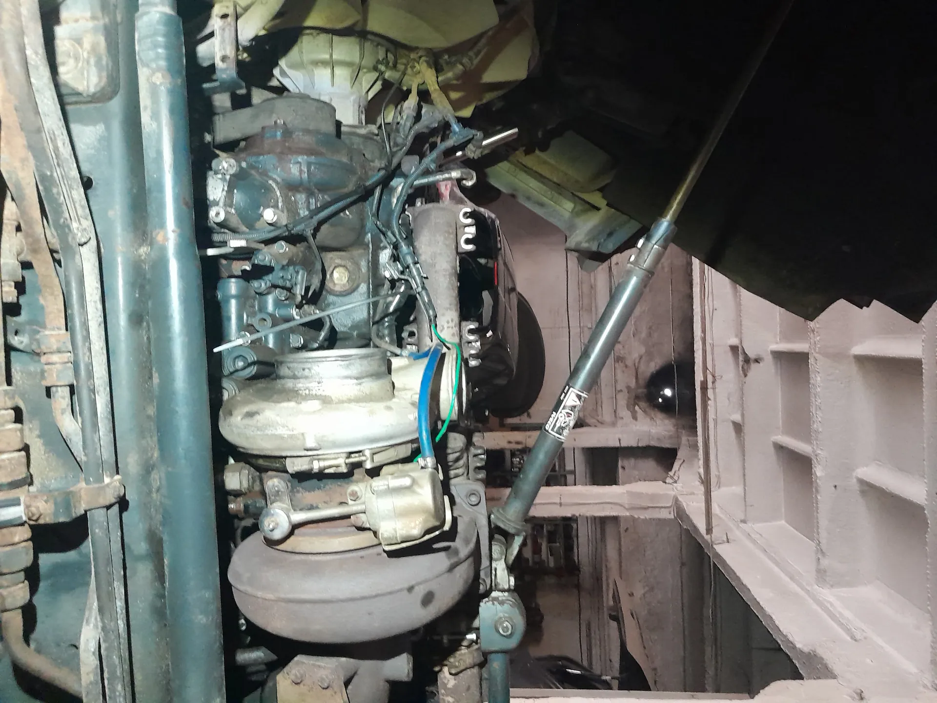 Zdjęcie przedstawia silnik pojazdu ciężarowego lub maszyny, widoczne są metalowe rury, przewody, turbodoładowacz oraz zardzewiałe elementy. W tle struktura betonowa garażu lub warsztatu z pionowymi belkami. Na jednej z rur znajduje się czarna etykieta z nieczytelnym białym tekstem. Oświetlenie jest ciemne, a całość wydaje się brudna i zużyta.
