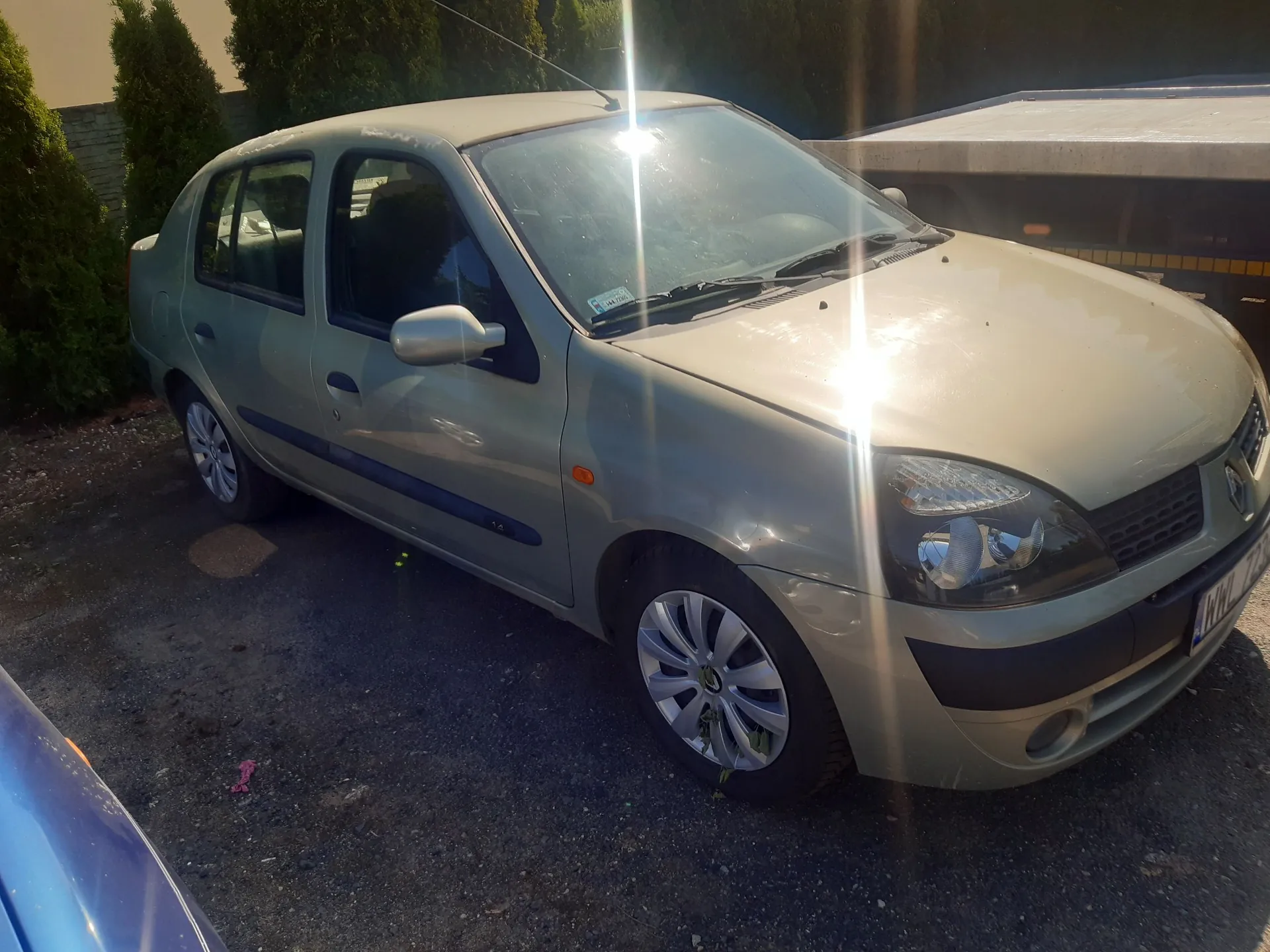 Srebrny samochód osobowy marki Renault Clio zaparkowany na podjeździe. Pojazd jest widoczny z przodu i z boku, w pełnym słońcu, które powoduje odblaski na masce i przedniej szybie. W tle widać zielone krzewy oraz fragment budynku.