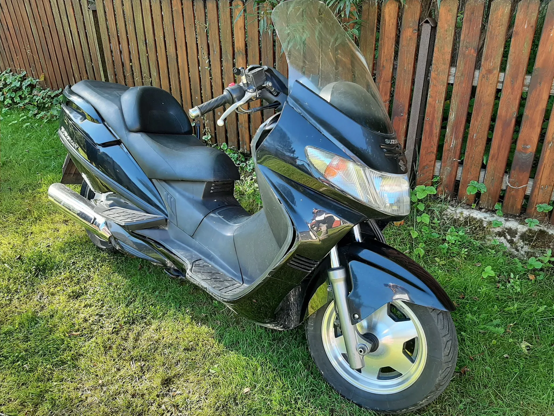Czarny skuter marki Suzuki zaparkowany na trawie na tle drewnianego ogrodzenia. Pojazd ma wysoką, przezroczystą szybę ochronną, srebrny wydech oraz srebrne felgi. Na boku skutera widoczny jest napis 'SUZUKI'.