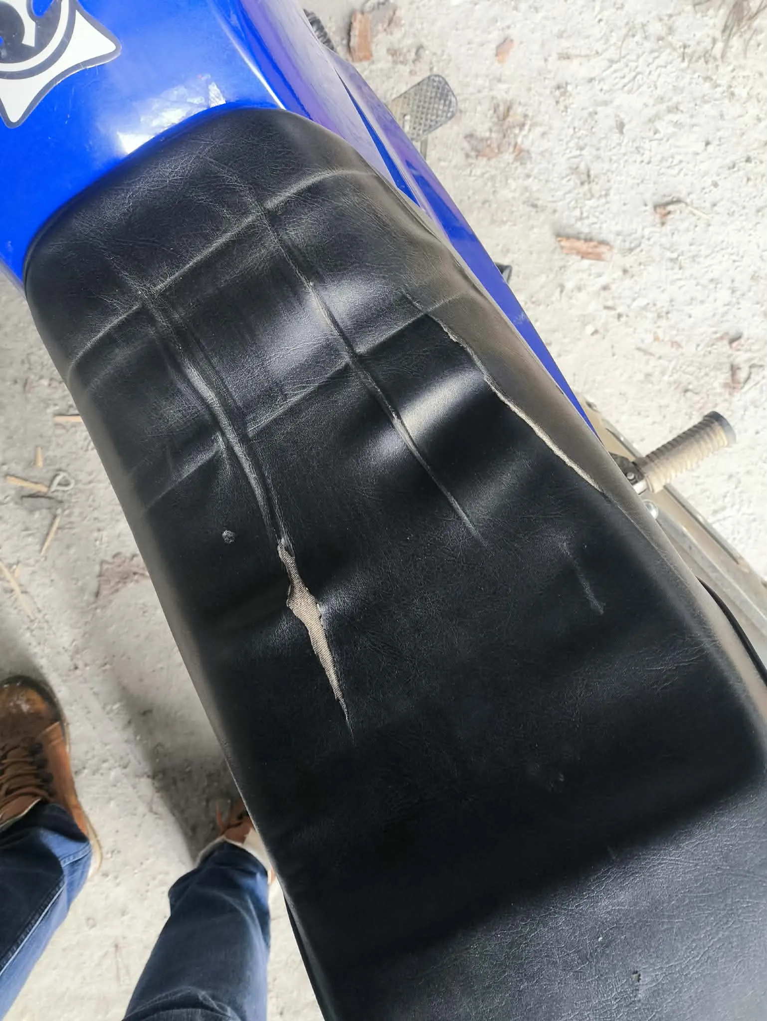 Czarny skórzany siedzenie motocykla z widoczną pionową rozdarcia, odsłaniającą jasny materiał pod spodem. Niebieska część motocykla z białym emblematem w lewym górnym rogu. W tle betonowa powierzchnia z drobnymi liśćmi i szczątkami. W lewym dolnym rogu widoczna część nogi osoby w niebieskich dżinsach i brązowych butach. Fragment kierownicy motocykla po prawej stronie.