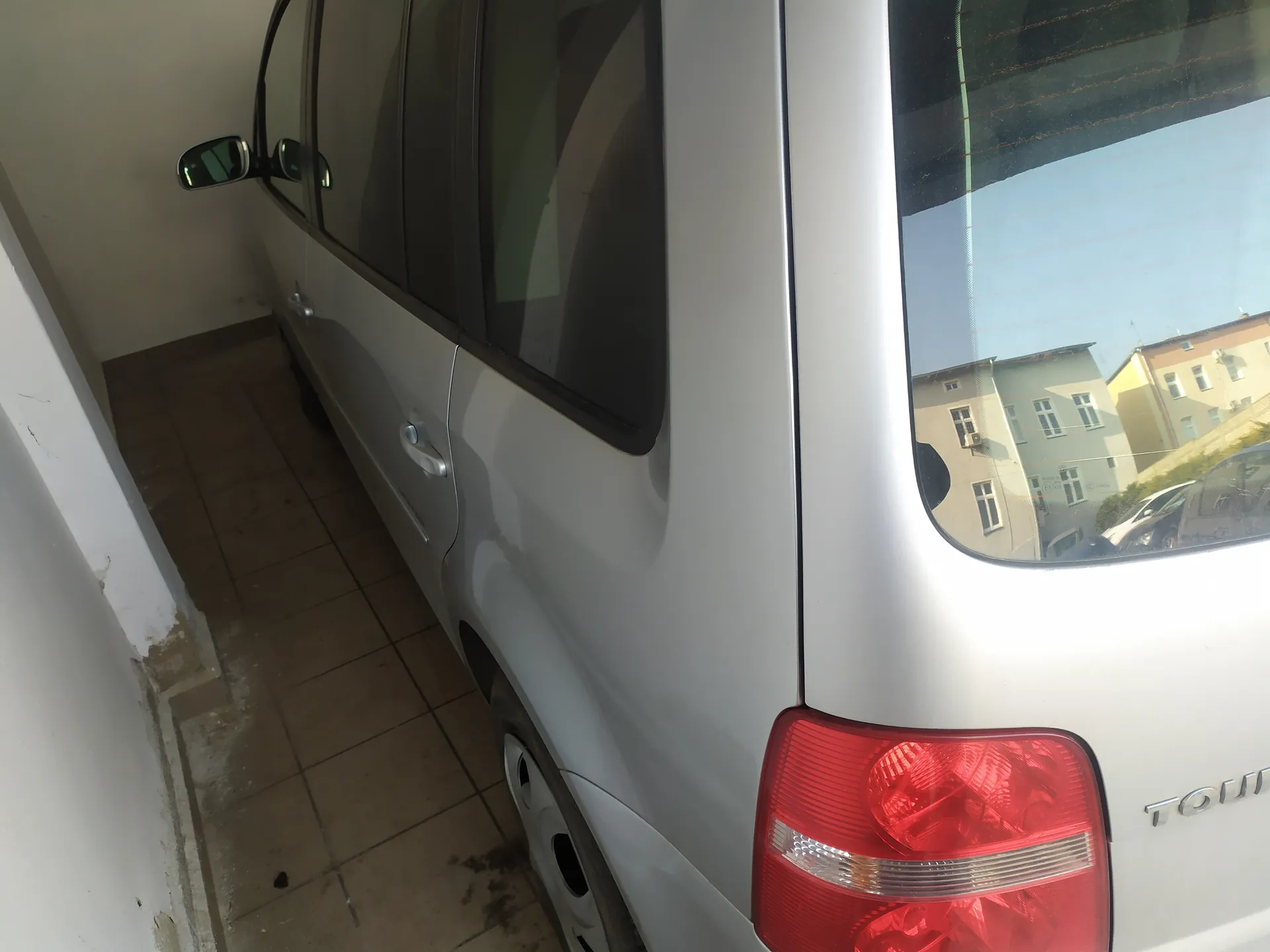 Srebrny samochód widziany z boku i tyłu, zaparkowany w zamkniętym pomieszczeniu z podłogą wyłożoną brązowymi płytkami. Widoczne są boczne lusterka, czerwone światło tylnego reflektora oraz fragment napisu 'TOUR' na tylnej części pojazdu. W tylnej szybie odbijają się budynki i inne samochody.