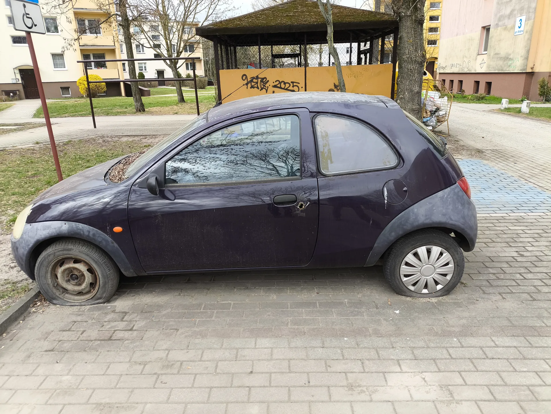 Mały, zniszczony fioletowy samochód osobowy zaparkowany na brukowanej powierzchni. Samochód ma uszkodzone przednie lewe koło, zarysowane karoserię i dziurę w drzwiach. W tle widoczne są bloki mieszkalne, drzewa bez liści, żółta bariera z graffiti zawierającym napis '2025', wózek na zakupy pełen śmieci oraz znak informujący o miejscu parkingowym dla osób niepełnosprawnych. Na budynku po prawej stronie znajduje się niebieski numer '3'.