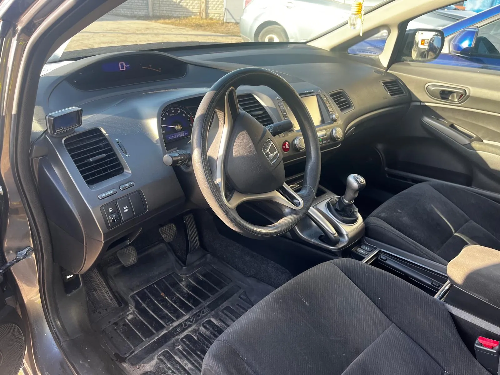 Wnętrze samochodu Honda Civic z widocznym kierownicą z logo Hondy, deska rozdzielcza z licznikami, przyciskami i ekranem, czarnymi siedzeniami, dźwignią zmiany biegów oraz dywanikiem z napisem "CIVIC". Drzwi kierowcy są otwarte, a na zewnątrz widoczne są inne zaparkowane pojazdy. Na wyświetlaczu deski rozdzielczej widoczna jest cyfra "0".