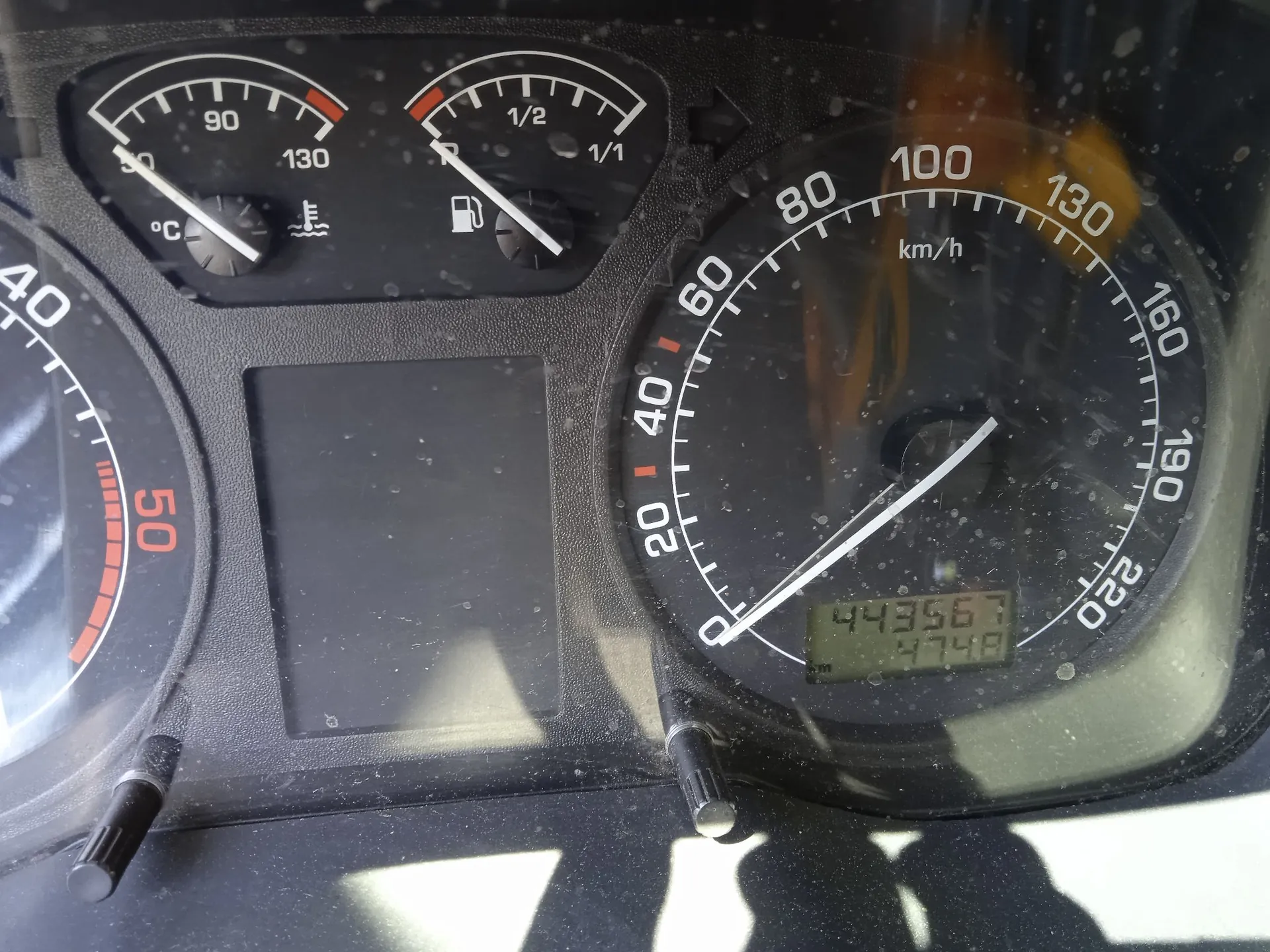 Zbliżenie na deskę rozdzielczą samochodu. Widoczny jest prędkościomierz z maksymalną wartością 220 km/h, wskazujący prędkość 0 km/h, oraz licznik kilometrów z wartością 44356,7. Po lewej stronie znajdują się wskaźniki temperatury cieczy chłodzącej oraz poziomu paliwa. Obudowa deski jest czarna i lekko zakurzona.