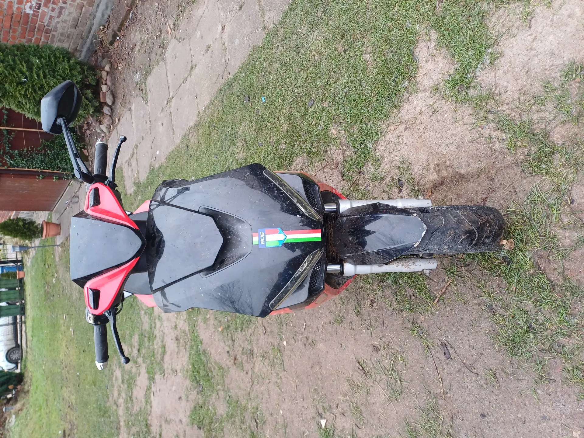 Motocykl czarno-czerwony z naklejką w kolorach włoskiej flagi i napisem "906" na czarnej części. Stoi na trawie i ziemi, w tle ceglana ściana, krzewy i chodnik. Widoczne lusterka, kierownica i przód pojazdu.