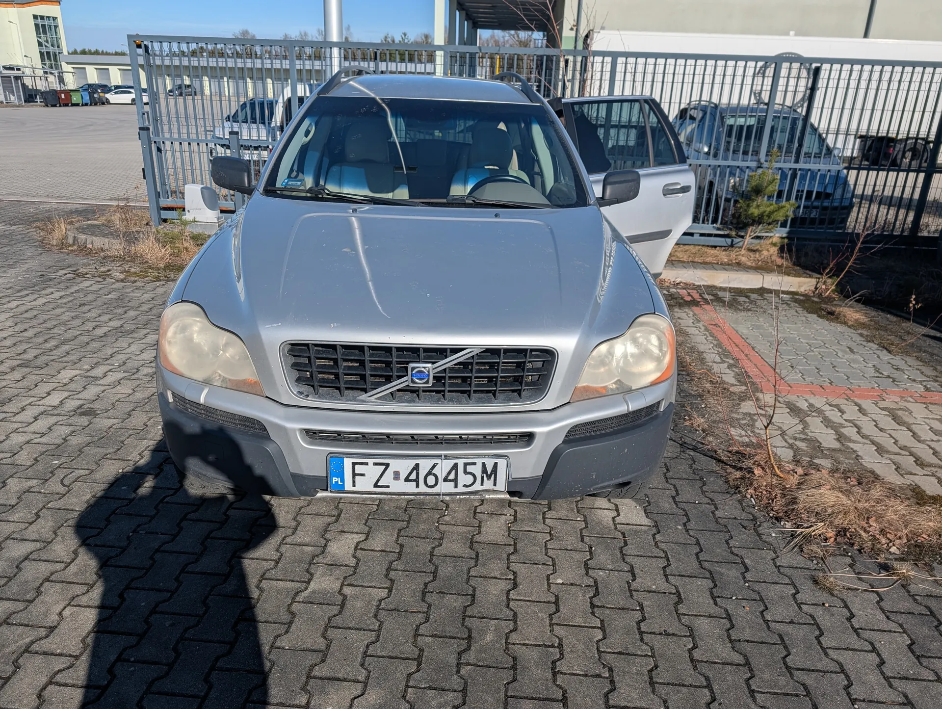 Srebrny samochód marki Volvo z tablicą rejestracyjną FZ 4645M (PL) zaparkowany na brukowanej powierzchni. Drzwi pasażera po prawej stronie są otwarte. W tle metalowe ogrodzenie, inne zaparkowane pojazdy, budynki oraz suche krzewy. Na tablicy rejestracyjnej widoczny jest niebieski pas z literą PL po lewej stronie.