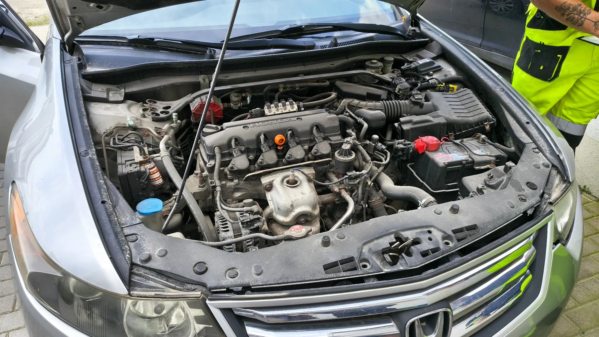 Widok z góry na otwartą maskę srebrnego samochodu marki Honda, ukazujący silnik spalinowy oraz elementy komory silnika, w tym akumulator po prawej stronie i zbiornik płynu spryskiwaczy po lewej. W prawym górnym rogu zdjęcia widoczny jest fragment osoby w jaskrawożółtej kamizelce odblaskowej.