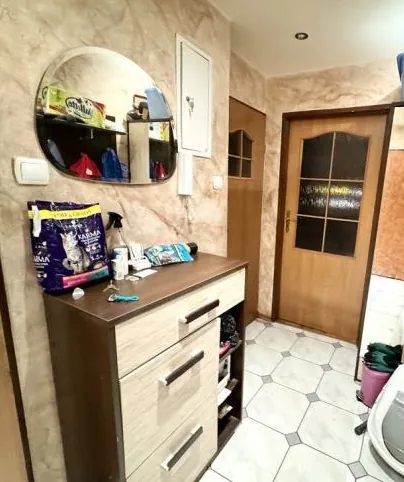 Wnętrze korytarza z drewnianą szafką o trzech szufladach i otwartym półkami po prawej stronie. Na szafce znajdują się: fioletowa torba z napisem 'KARMA', spray, biały kubek, niebieski przedmiot i inne drobne elementy. Nad szafką wiszy okrągłe lustro odbijające półki z kolorowymi torbami i pudełkami. Ściany wykończone są wzorzystą tapetą w odcieniach beżu. Po prawej stronie widoczne są dwa drewniane drzwi z szybkami, jedne z nich prowadzą do innego pomieszczenia. Podłoga wyłożona białymi płytkami z geometrycznym wzorem. W prawym dolnym rogu stoi różowy wiadro z zielonym ręcznikiem. Na suficie znajduje się okrągłe oświetlenie.