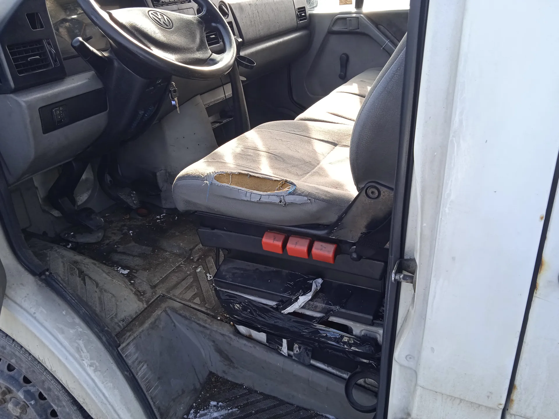 Wnętrze samochodu marki Volkswagen z widocznym kierownicą z logo VW, uszkodzonym siedzeniem kierowcy z odsłoniętą pianką, trzema czerwonymi przyciskami na mechanizmie siedzenia, brudną podłogą z resztkami śniegu i kluczami w zapłonie. Drzwi samochodu są otwarte, a zewnętrzna część nadwozia jest biała. W tle widoczne są elementy deski rozdzielczej i klimatyzacji.