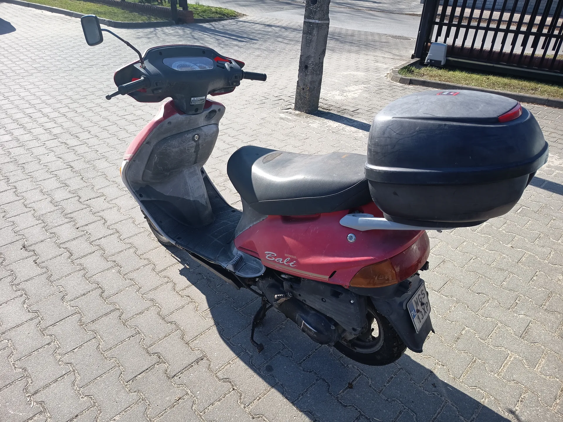 Czerwony skuter marki Bali z czarnym siedzeniem i czarnym bagażnikiem na tylnej części. Skuter stoi na brukowanej powierzchni. Widoczna tablica rejestracyjna z napisem HGS 13XP. W tle drzewo, metalowe ogrodzenie i trawnik. Słoneczny dzień, cienie wskazują na jasne oświetlenie.