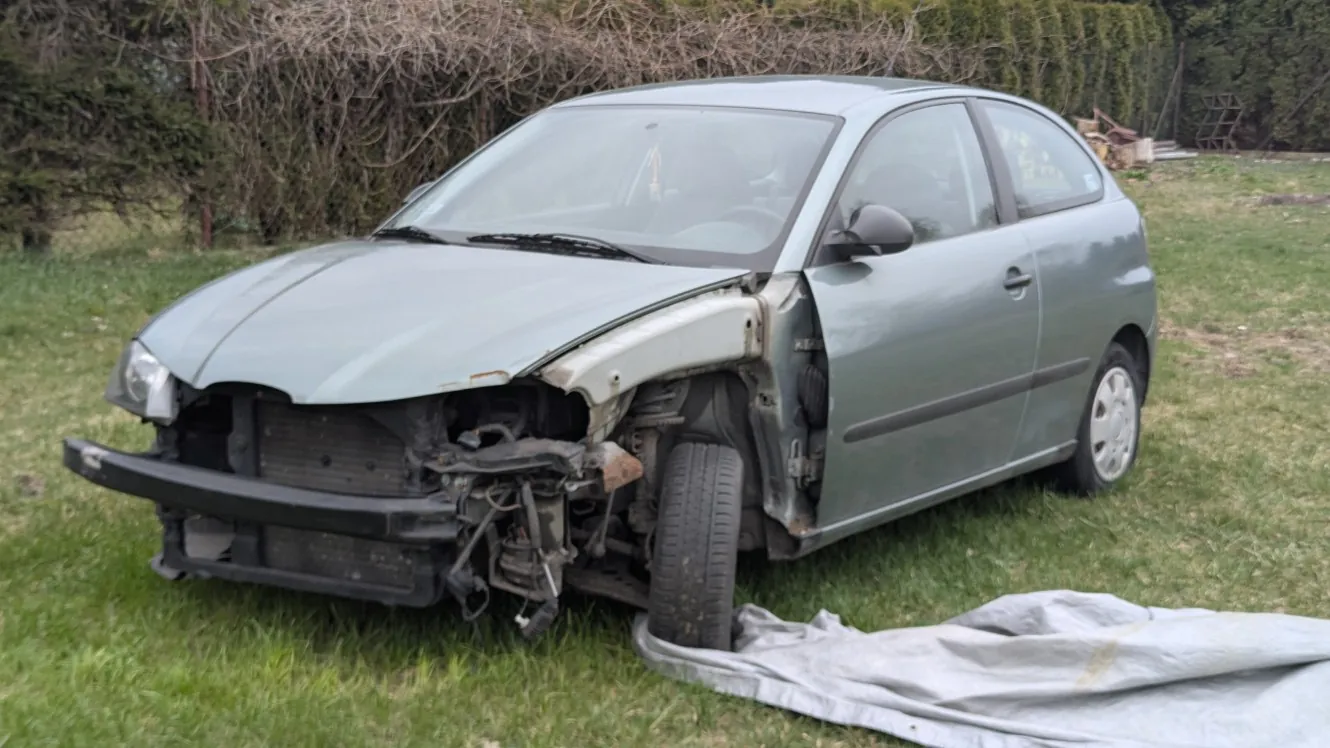 Uszkodzony jasnozielony samochód osobowy typu hatchback z zniszczonym przodem, stojący na trawie. Widoczne uszkodzenia obejmują zdeformowaną maskę, brak zderzaka oraz odsłonięte elementy podwozia. W tle krzewy i zarośla, na ziemi leży szary płacht tkaniny.