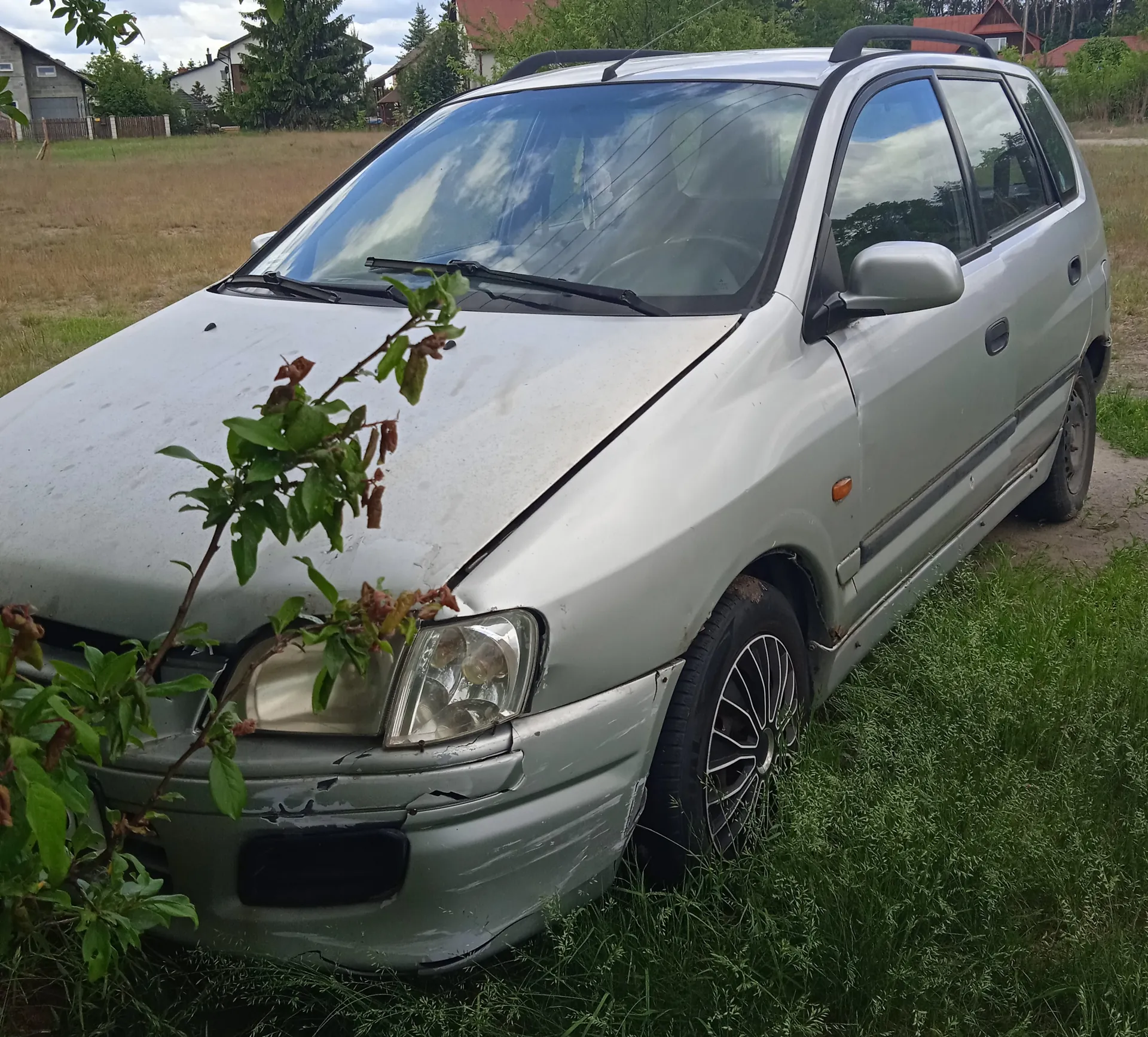Srebrny samochód osobowy typu kombi z widocznymi uszkodzeniami na przednim zderzaku i karoserii, zaparkowany na trawie. W tle pola, domy, drzewa oraz częściowo zachmurzone niebo. Przed samochodem rośnie gałązka z zielonymi liśćmi. Opony mają metalowe felgi, a na dachu widoczne są uchwyty dachowe.