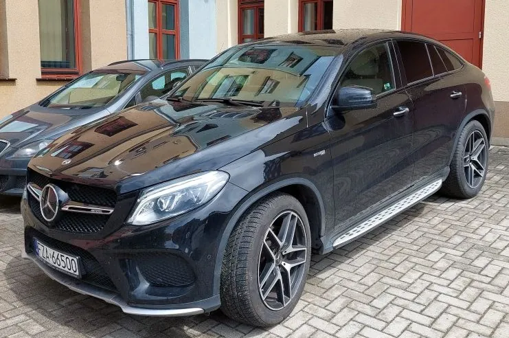 Czarny samochód Mercedes-Benz GLE Coupe zaparkowany na brukowanej powierzchni. Widoczna tablica rejestracyjna F LA 66500. W tle szary samochód oraz budynek z czerwonymi ramami okiennymi i czerwonymi drzwiami.