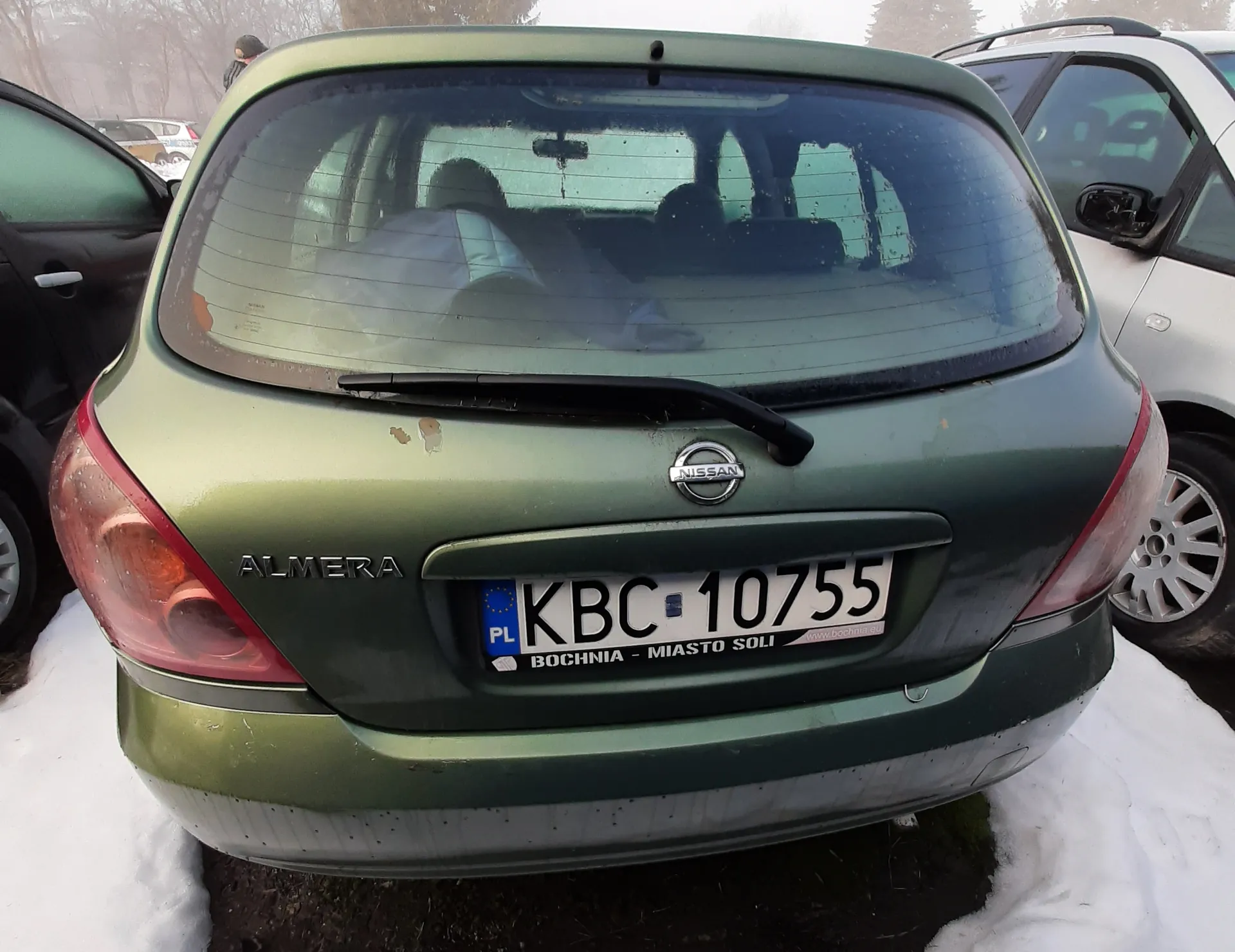 Z tyłu zielony samochód Nissan Almera z tablicą rejestracyjną KBC 10755. Na tablicy widoczny napis PL oraz naklejka z napisem BOCHNIA - MIASTO SOLI. Samochód stoi na śniegu, w tle inne pojazdy i drzewa. Na tylnym szybie widać sylwetki osób wewnątrz pojazdu.
