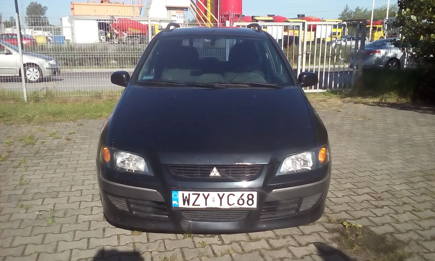 Czarny samochód osobowy marki Mitsubishi zaparkowany przodem do obiektywu na kostce brukowej. Pojazd posiada polską tablicę rejestracyjną o numerze WZY YC68. W tle widoczny jest metalowy płot, inne zaparkowane samochody oraz czerwone zbiorniki przemysłowe.