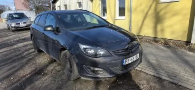 Czarny samochód marki Opel z tablicą rejestracyjną P12515 zaparkowany na ulicy. W tle widoczny srebrny samochód oraz żółty budynek z oknami. Na jezdni brudne plamy i cienie drzew.