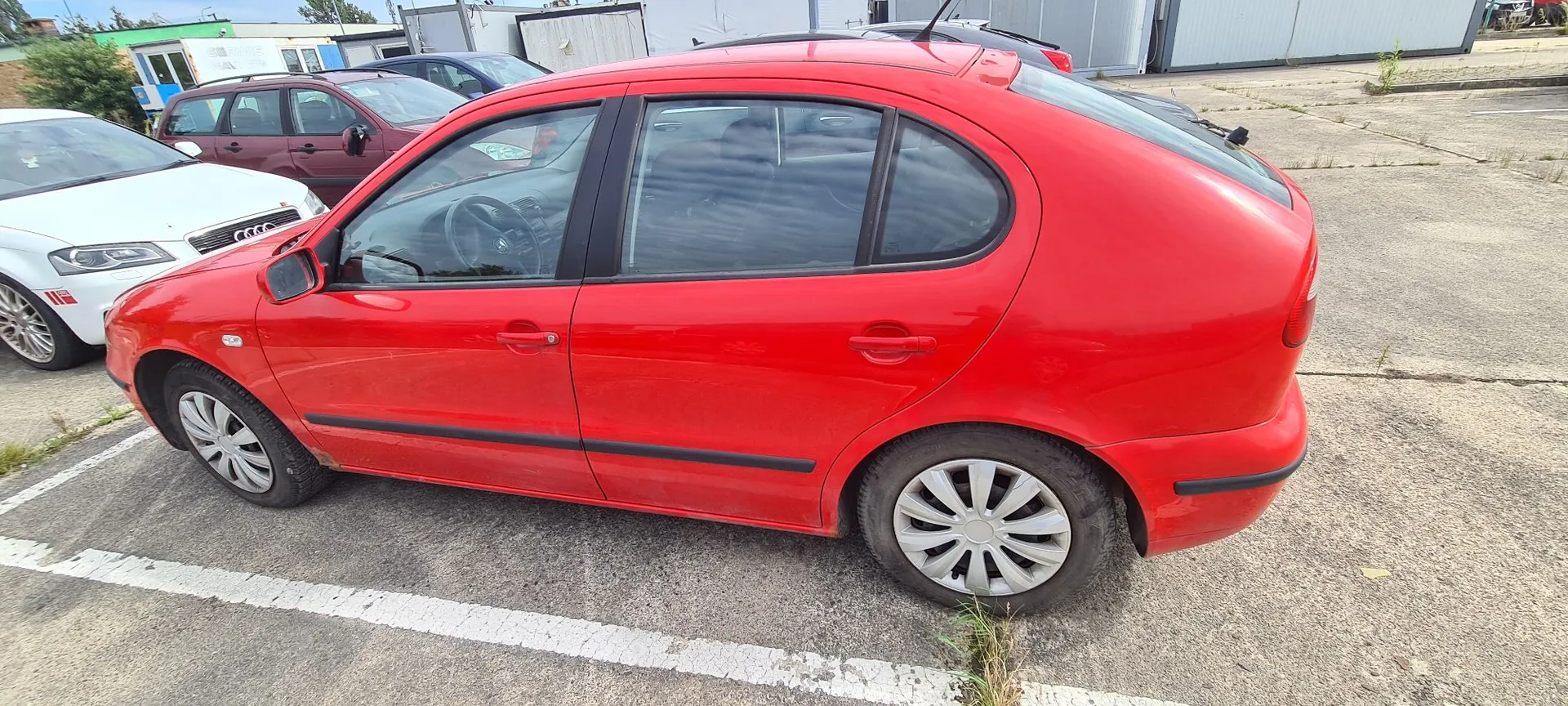 Profil czerwonego samochodu osobowego typu hatchback zaparkowanego na betonowym placu. W tle widoczne są inne samochody: biały po lewej stronie oraz bordowy i niebieski za czerwonym autem. W oddali znajdują się budynki przemysłowe.