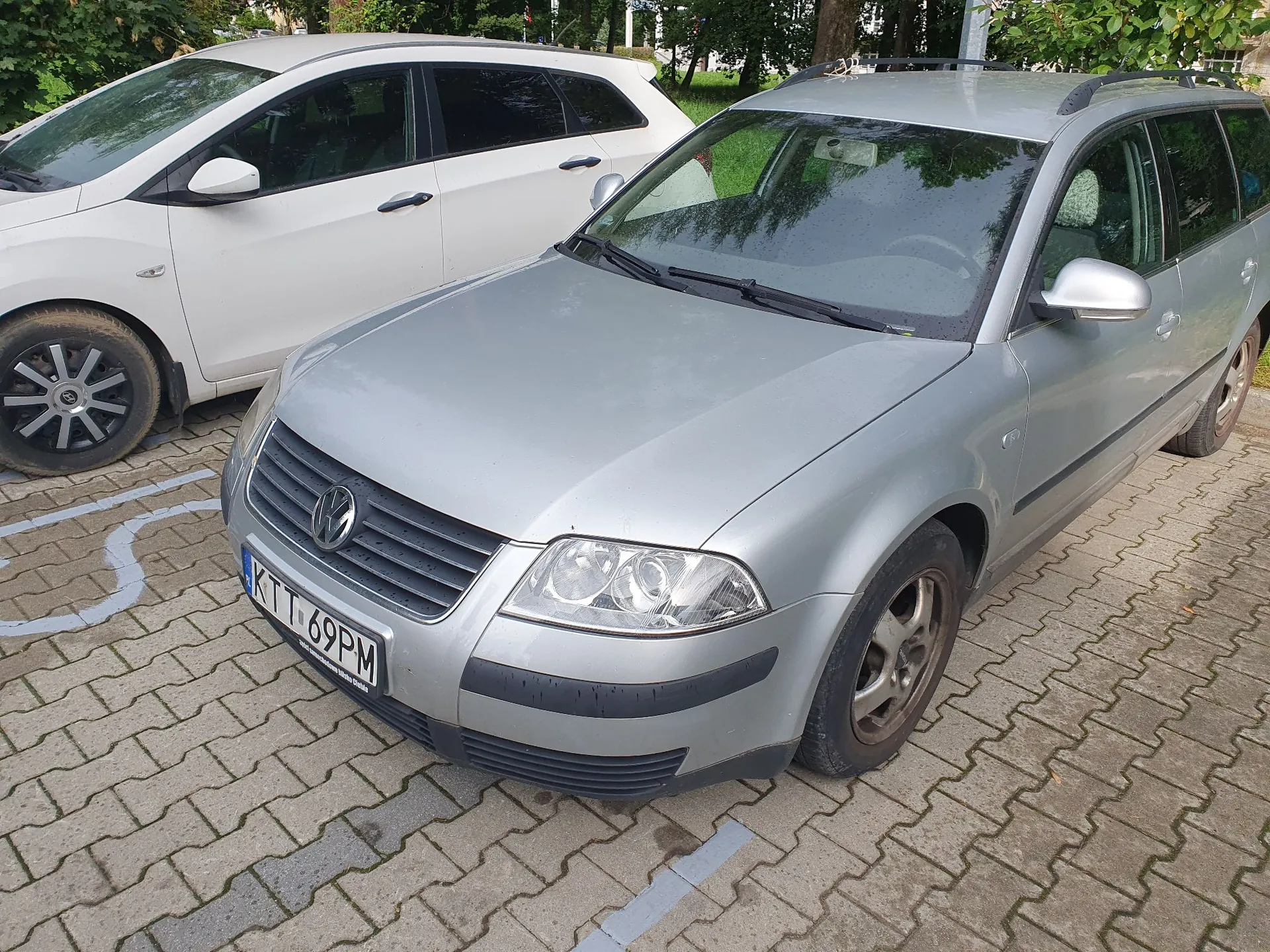 Srebrny samochód marki Volkswagen w nadwoziu kombi zaparkowany na kostce brukowej. Obok niego stoi biały samochód. Srebrny samochód ma tablicę rejestracyjną o numerze KTT 69PM.