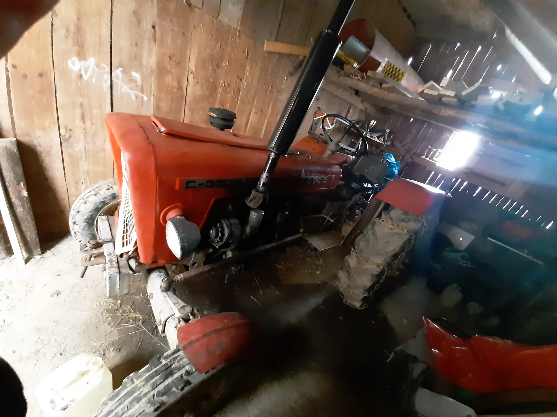Czerwony traktor model C-330 w stodole z drewnianymi ścianami. Na traktorze widoczny napis C-330. W tle narzędzia, półki z przedmiotami i otwór wpuszczający światło. Na podłodze biały pojemnik i ślad po wodzie. Ściany pokryte białymi napisami.
