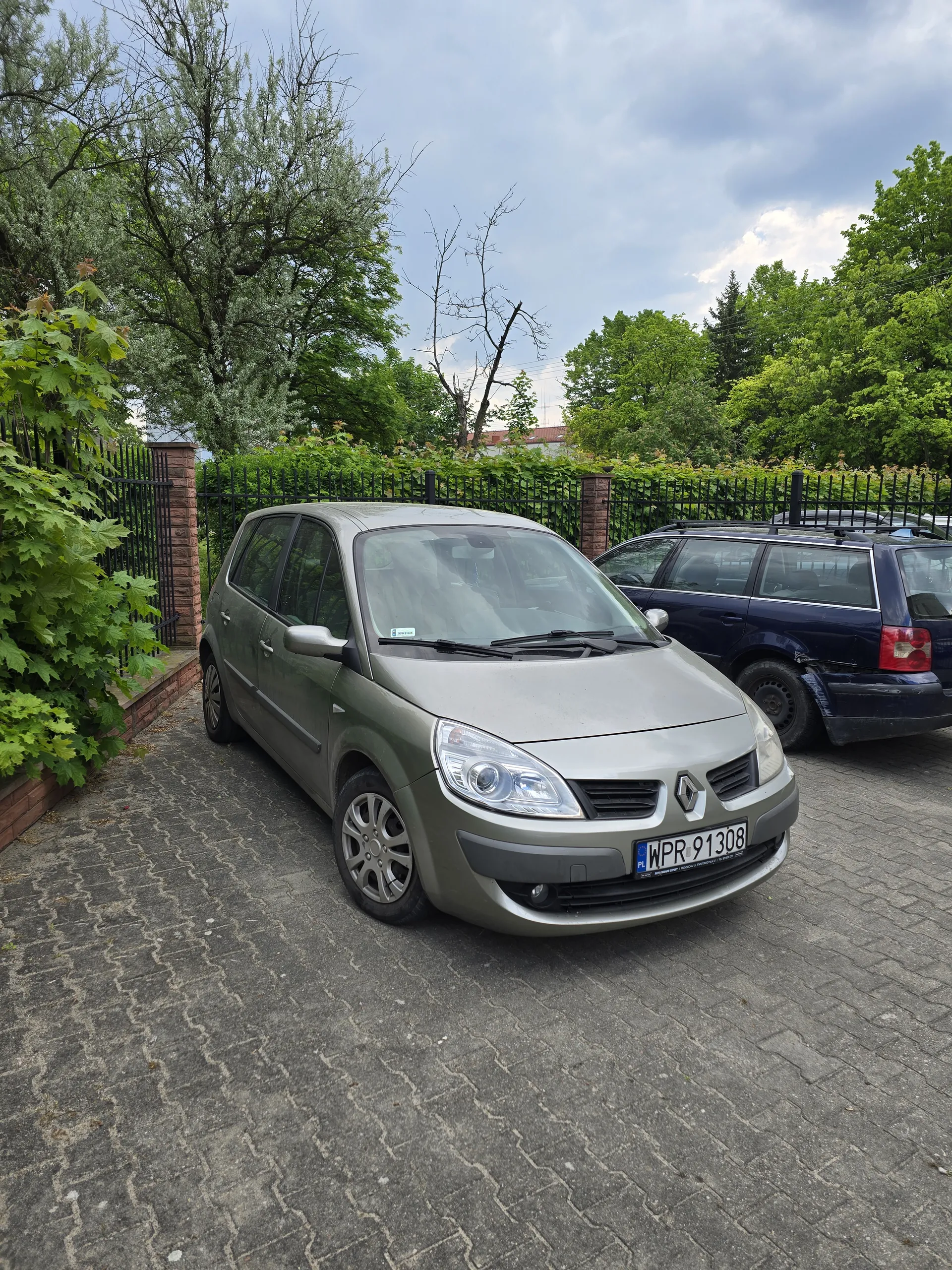 Dwa zaparkowane samochody na brukowanej powierzchni. W pierwszym planie srebrny samochód marki Renault z tablicą rejestracyjną WPR 91308. Za nim ciemnoniebieski samochód. W tle zielone drzewa, czarne metalowe ogrodzenie, ceglany mur i chmurne niebo. Na pierwszym planie widoczne są również zielone krzewy i liście.