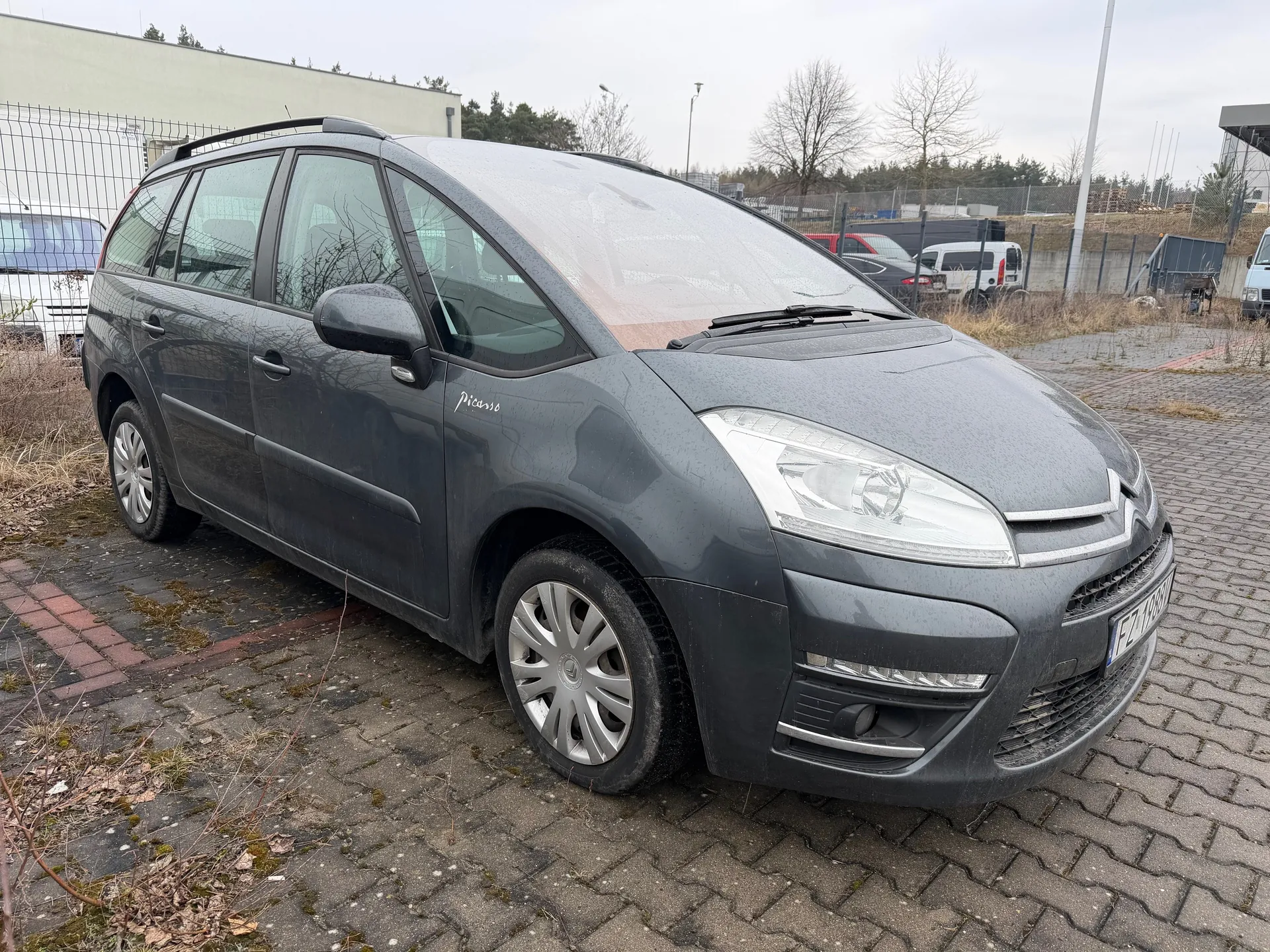 Szary samochód Citroën Picasso zaparkowany na brukowanej powierzchni. Po lewej stronie widoczne są suche krzewy i fragment ogrodzenia, w tle budynek, drzewa oraz inne pojazdy. Na boku pojazdu widnieje napis 'Picasso', a na przodzie tablica rejestracyjna z częściowo czytelnym numerem 'FZ 1987H'. Samochód posiada dachowe pręty i srebrne felgi. Niebo jest zachmurzone.