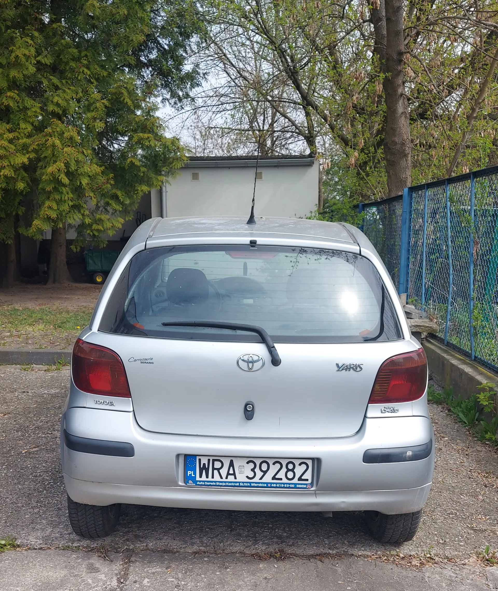Tylny widok srebrnego samochodu Toyota Yaris zaparkowanego na żwirowym podjeździe. Na tablicy rejestracyjnej widoczny jest napis PL WRA 39282. Po lewej stronie tylnego pasa widnieje napis Carrosserie Serrano, a po prawej Yaris. W tle drzewa, niebieskie ogrodzenie siatkowe oraz budynek. Samochód ma czarny wycieraczka na tylnej szybie i antenę na dachu.