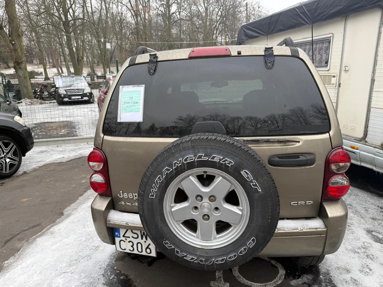 Z tyłu widok brązowego Jeepa 4x4 CRD z zamontowanym na bagażniku zapasowym kołem oznaczonym 'Wrangler ST' i 'Goodyear'. Tablica rejestracyjna PL ZSW C306. Na szybie tylnej naklejka z napisem 'SR ZSW C306'. W tle inne pojazdy, namiot, drzewa bez liści oraz śnieg na ziemi. Parkowanie na otwartym obszarze z widocznymi śladami zimy.