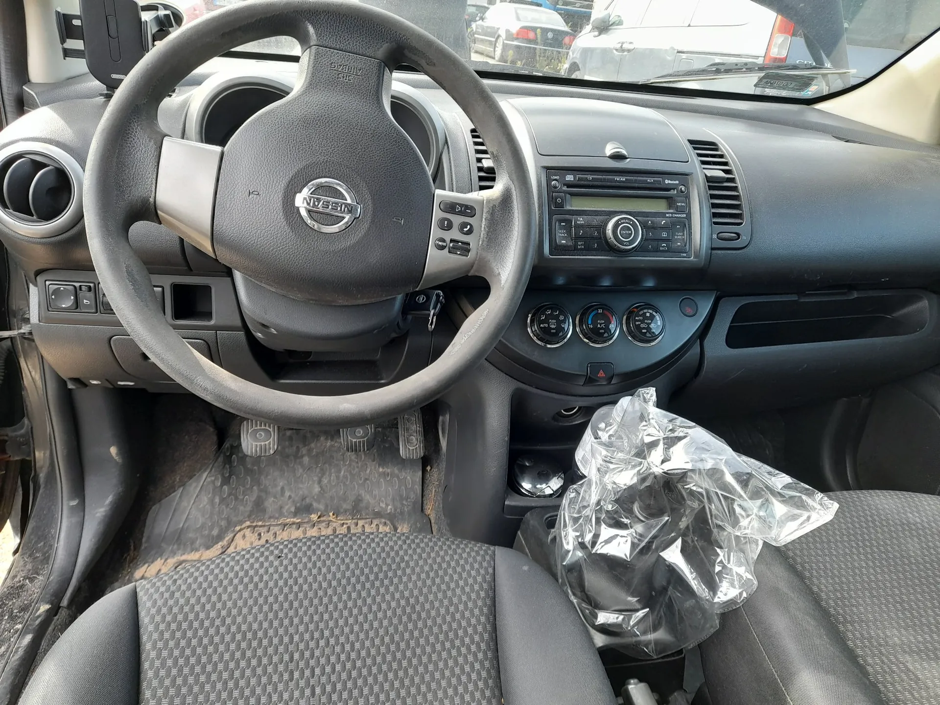 Wnętrze samochodu marki Nissan widziane z perspektywy kierowcy. Widoczna jest czarna kierownica z logo Nissan na środku, deska rozdzielcza z radiem, pokrętłami klimatyzacji oraz schowkiem. Na siedzeniu pasażera leży pognieciona, przezroczysta foliowa torba. Podłoga wyłożona jest gumowymi dywanikami.