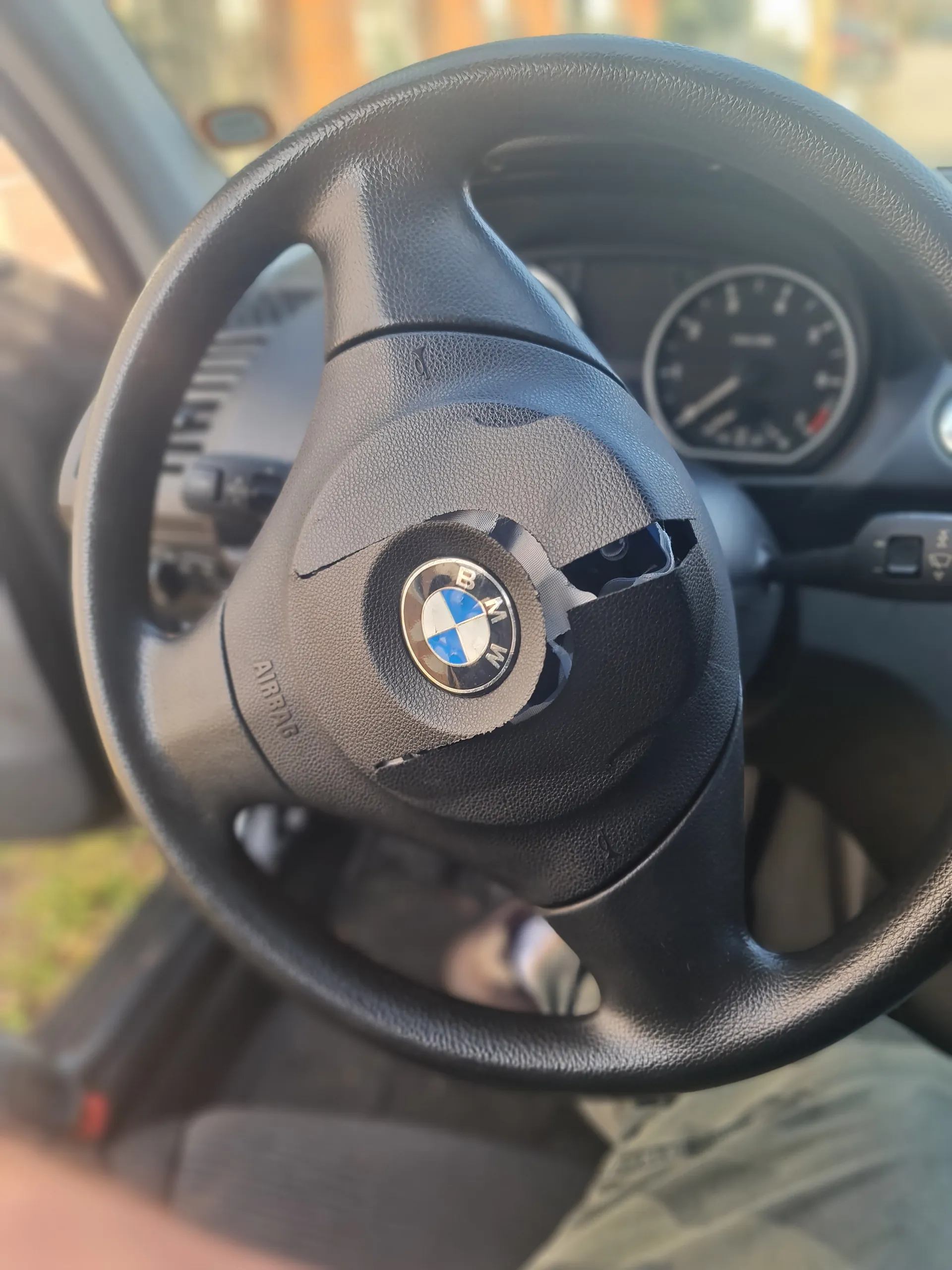 Czarna kierownica samochodu marki BMW z widocznym logo w centrum. Na kierownicy widoczne uszkodzenie – rozerwana obudowa w górnej części. Poniżej logo napis 'AIRBAG'. W tle zegar prędkościowy, dźwignia zmiany biegów oraz fragment nogi osoby w kamuflażowych spodniach. Drzwi samochodu są otwarte, a na zewnątrz rozmyty widok zieleni i budynku.