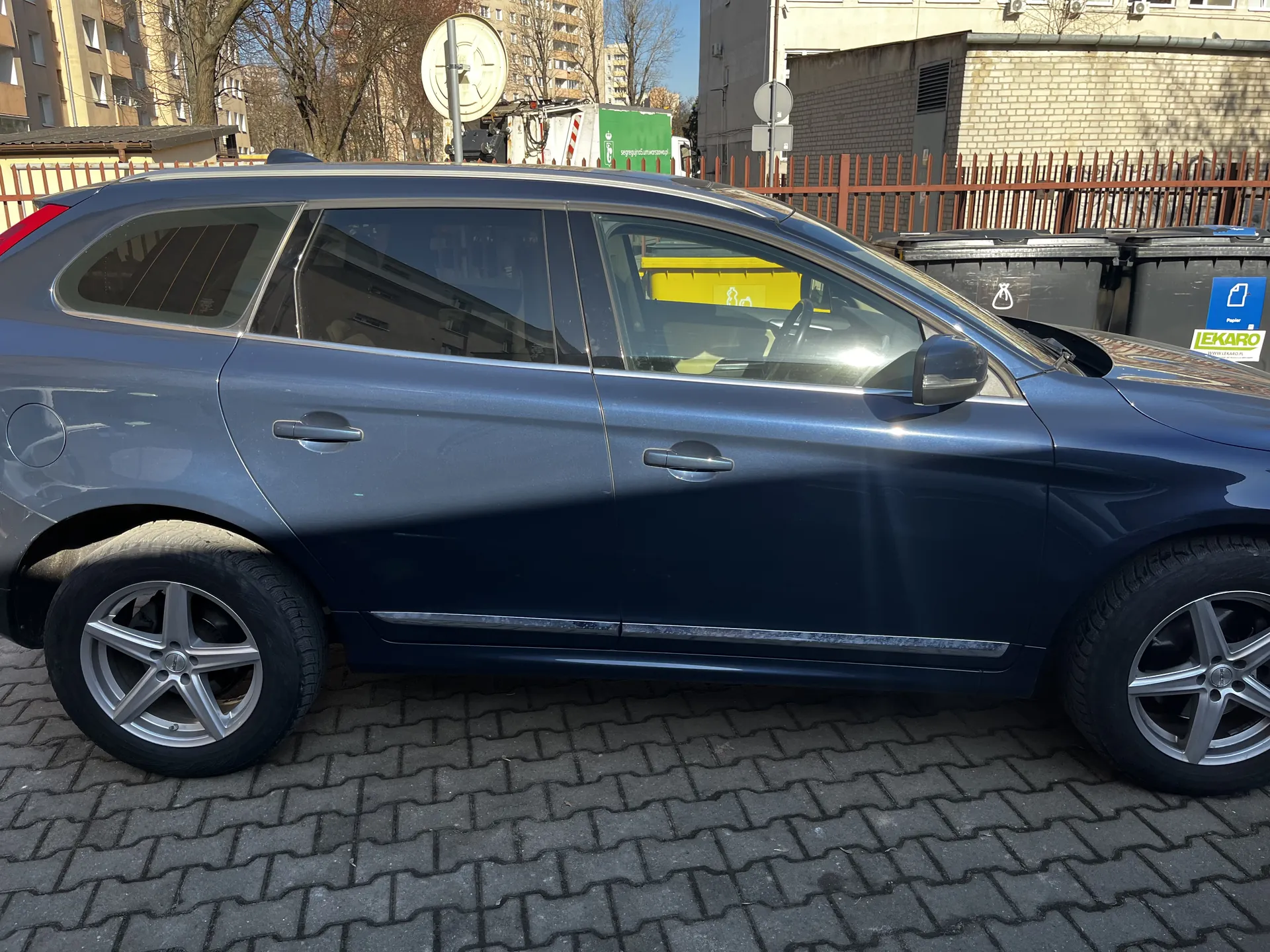 Błękitny samochód typu SUV zaparkowany na brukowanej powierzchni. Widoczna boczna strona pojazdu z srebrnymi felgami, czarnymi oponami i chromowanymi uchwytami drzwiowymi. W tle budynki mieszkalne, czerwone ogrodzenie, drzewa bez liści, zielony pojazd ciężarowy oraz czarne pojemniki na odpady. Na jednym z pojemników widoczny niebieski znak z napisem "LEKARO" oraz biały symbol. Słoneczna pogoda, cień pada na samochód i bruk.