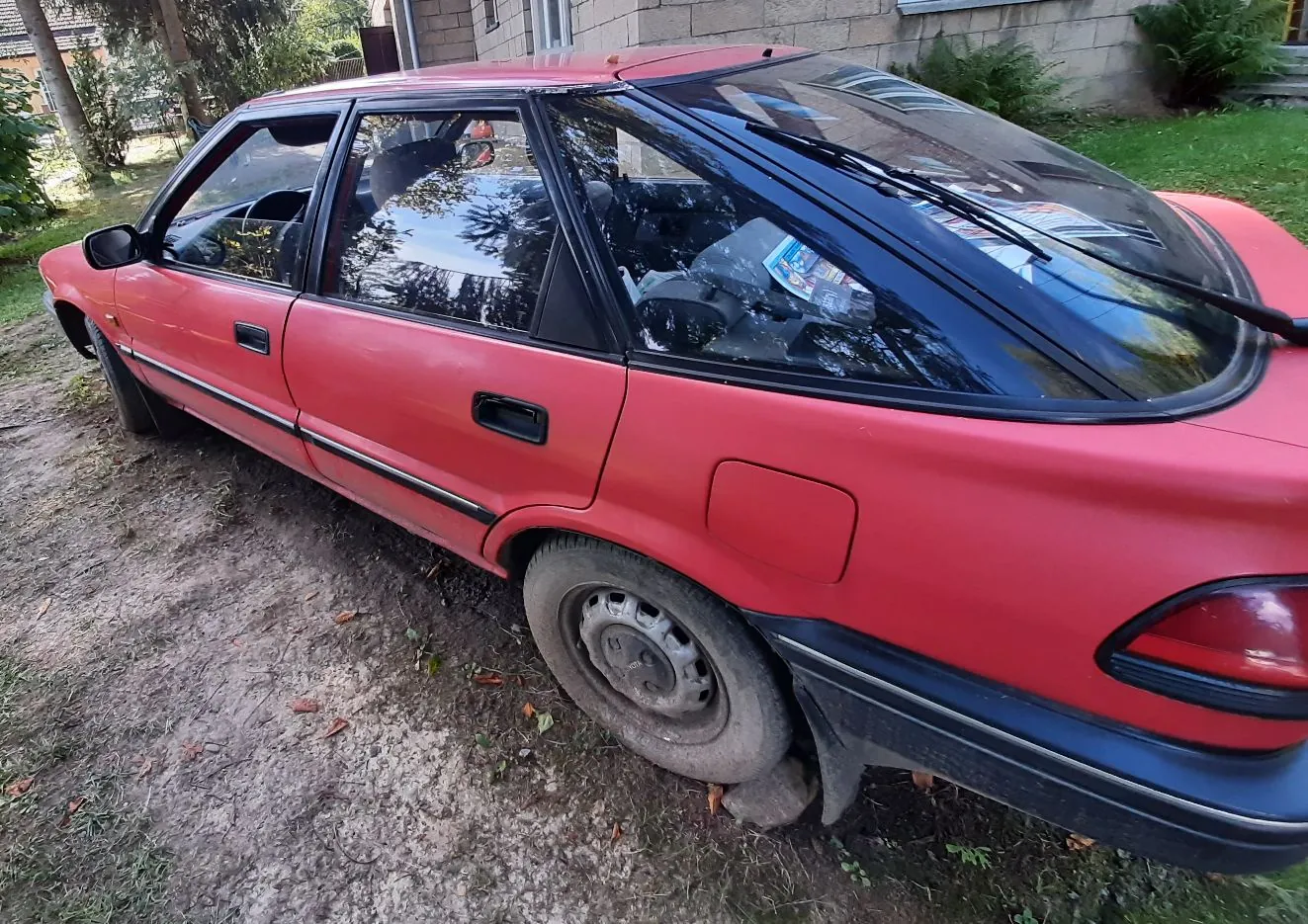 Czerwony samochód osobowy typu hatchback zaparkowany na piaszczystym podjeździe obok trawnika i budynku. Widok z boku i z tyłu pojazdu.